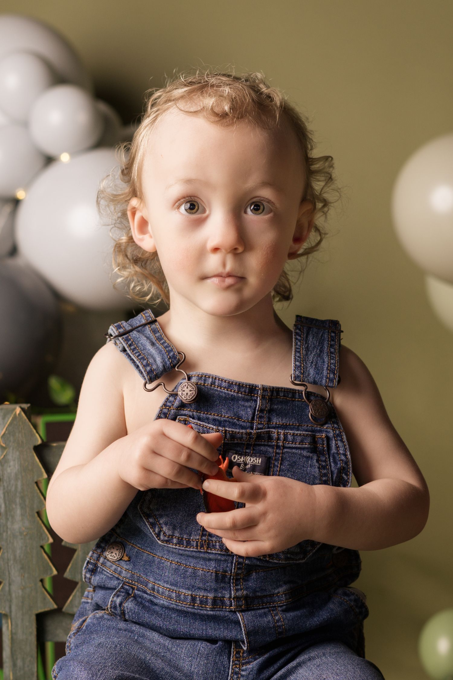 A small child in blue jean overalls poses against a tan backdrop with white balloons.