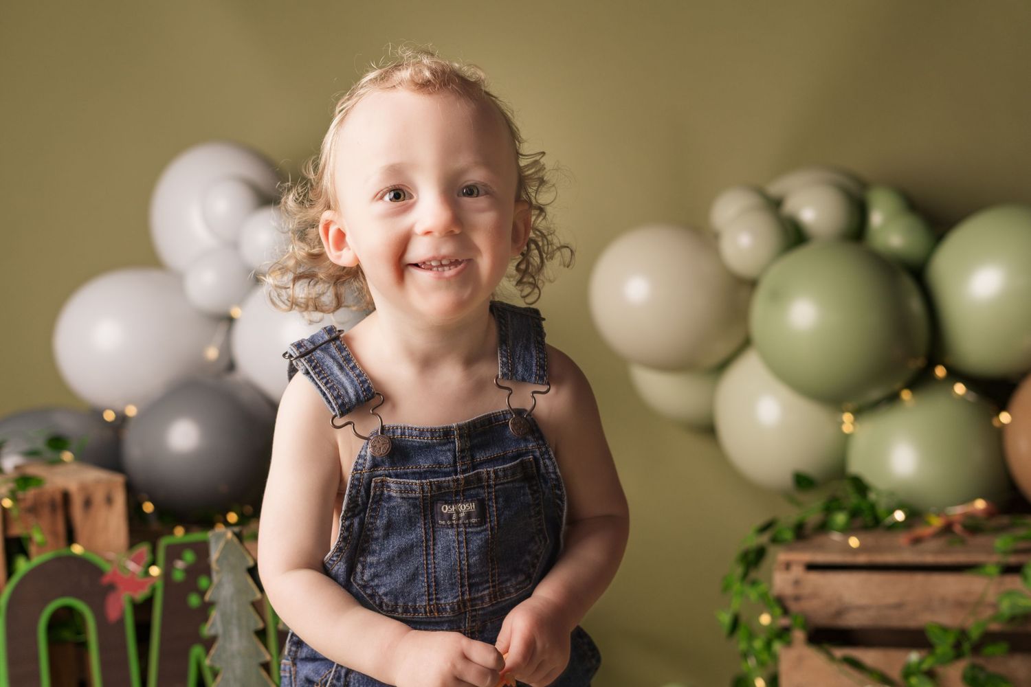 Child in denim overalls sits on wooden floor with green and white balloons and string lights in background.