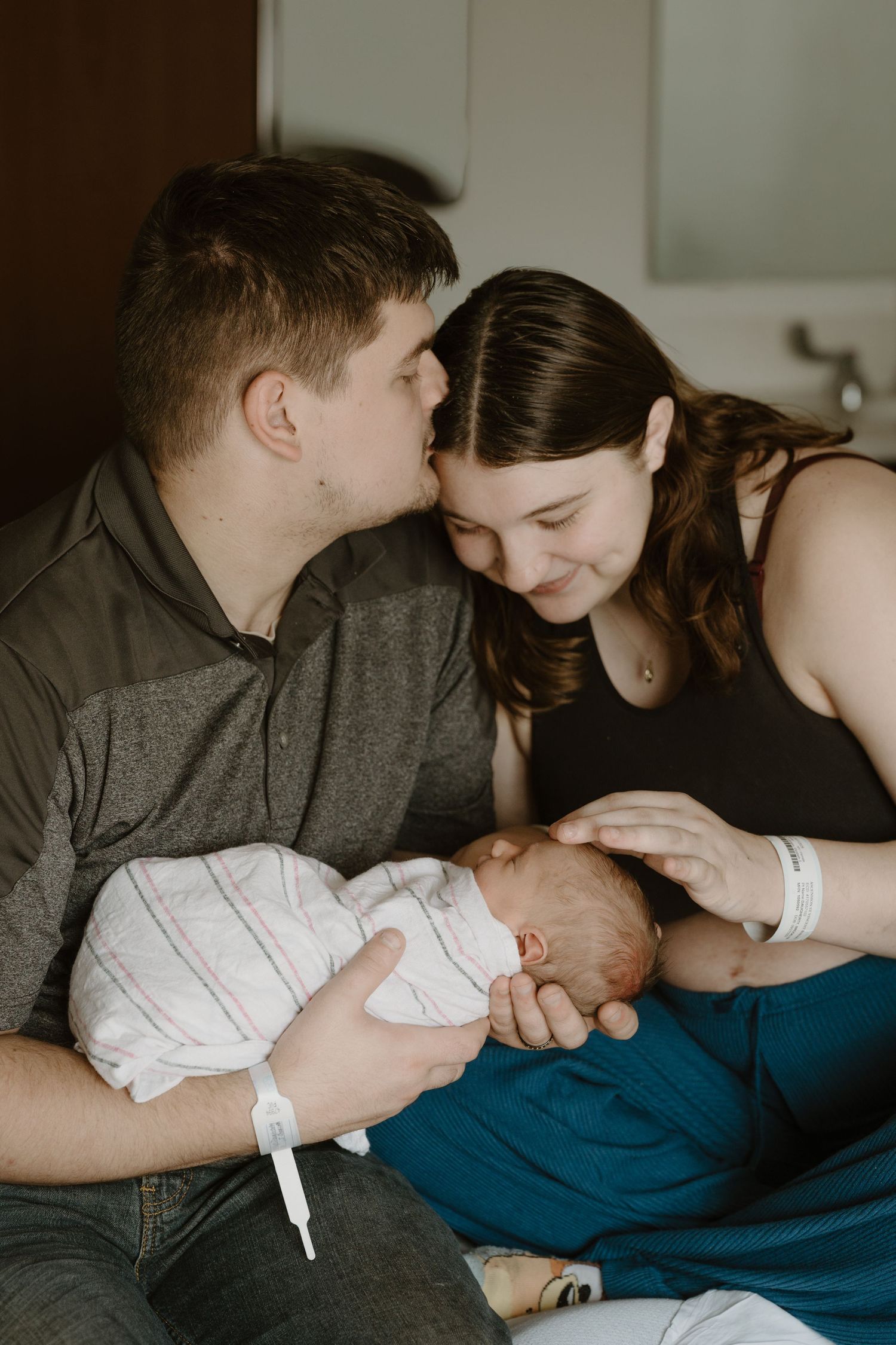 New parents share a tender moment kissing over their swaddled newborn baby during Fresh 48 in-hospital newborn photography session Indiana