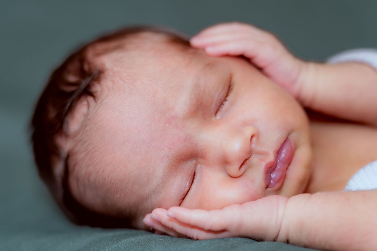 Sleeping newborn baby with dark hair rests peacefully against white bedding in soft natural light.