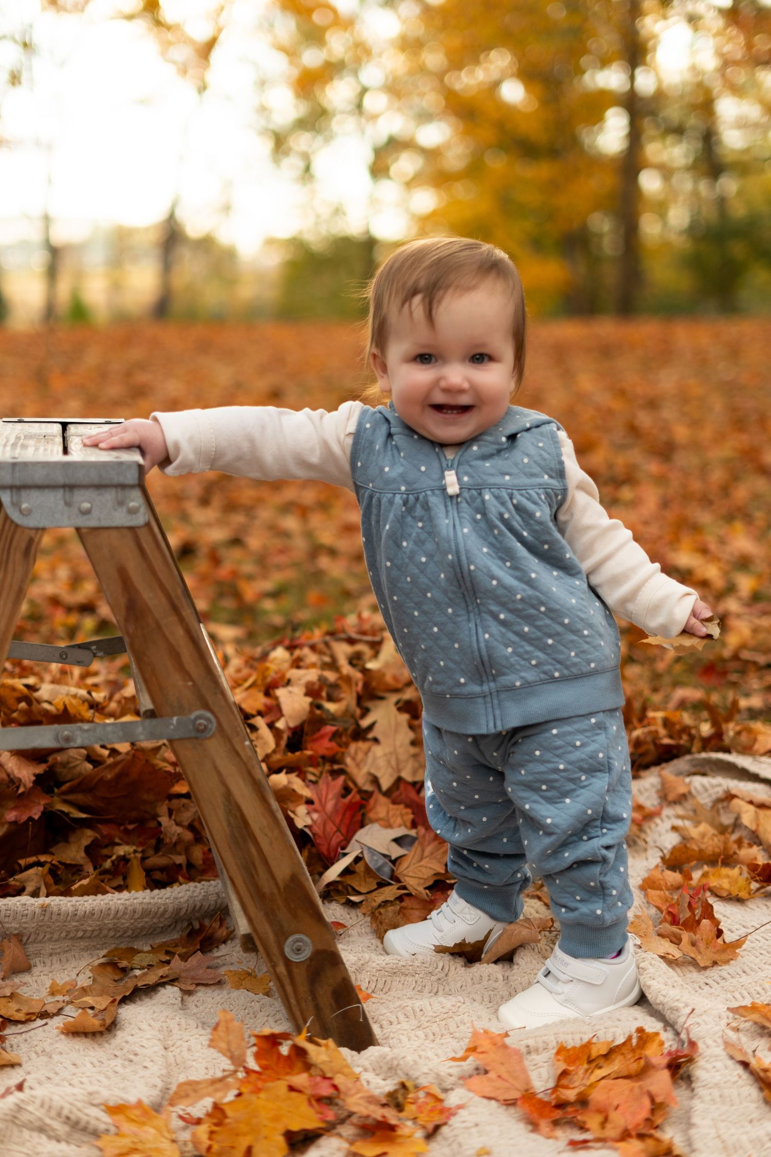 Toddler in denim outfit exploring fall leaves on white blanket during golden hour.