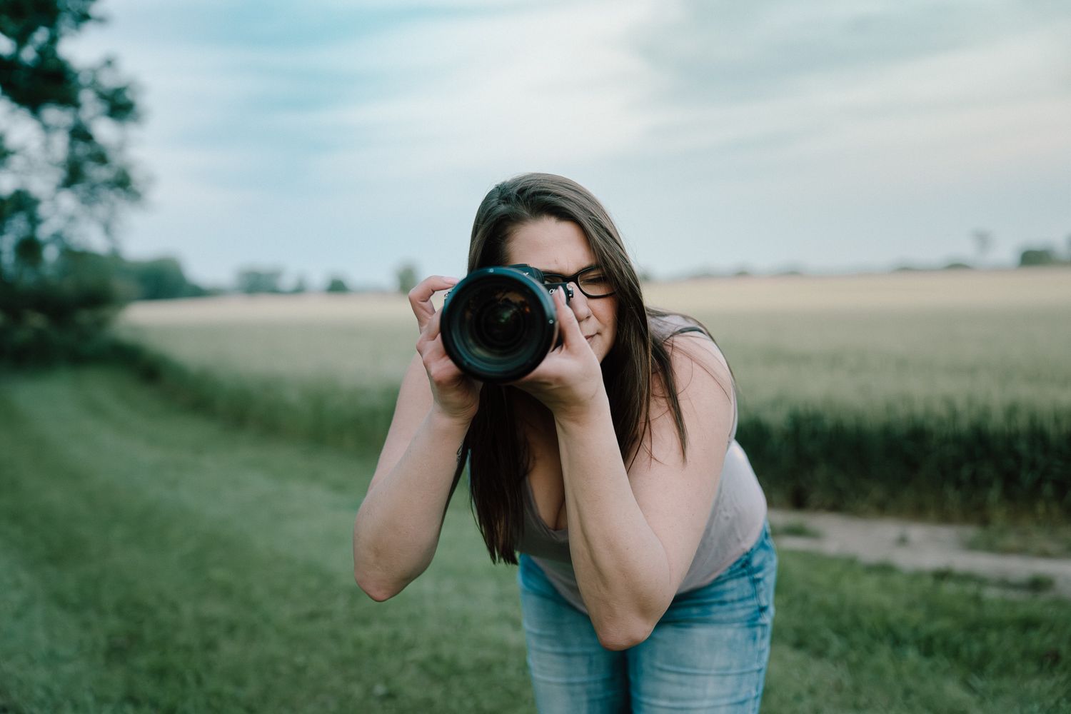 Woman with glasses photographing outdoors with a DSLR camera in a green field under a cloudy sky.