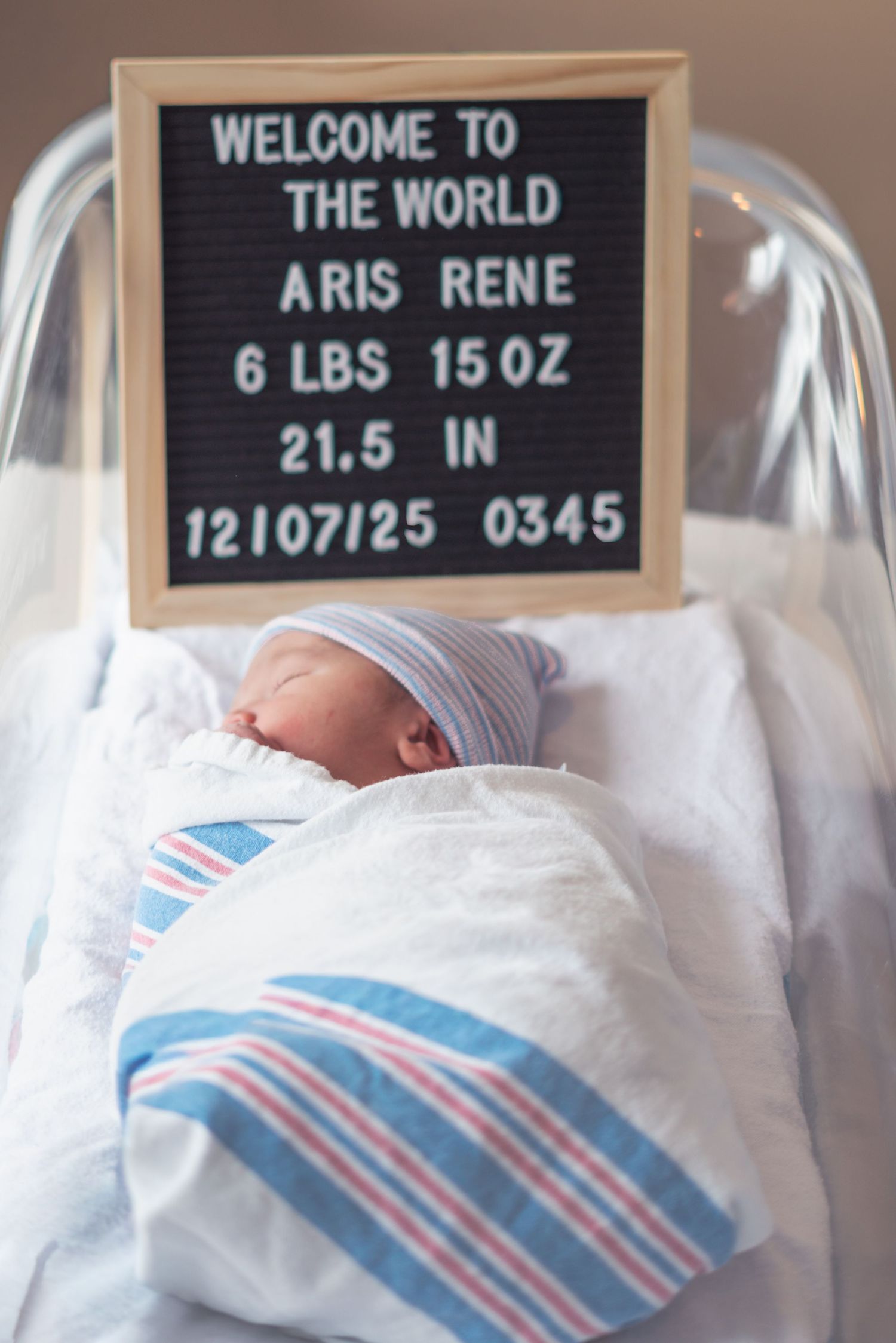 Newborn baby in hospital bassinet with blue and pink striped blanket under welcome sign and birth statistics.