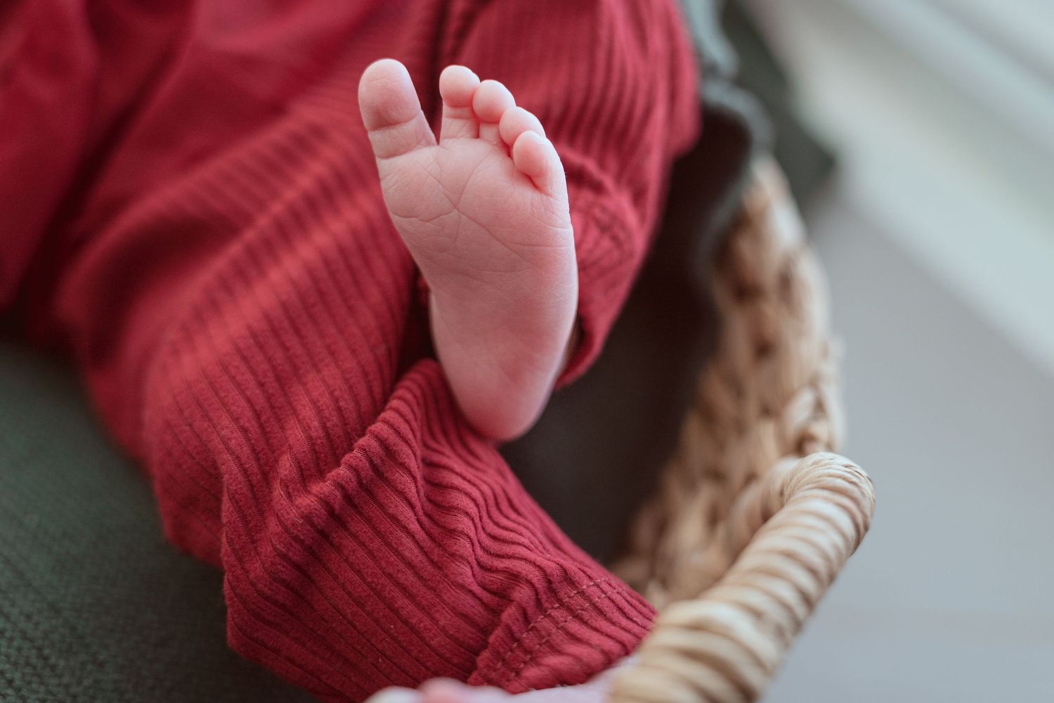 A tiny baby foot in red knit clothing rests on a woven basket edge.