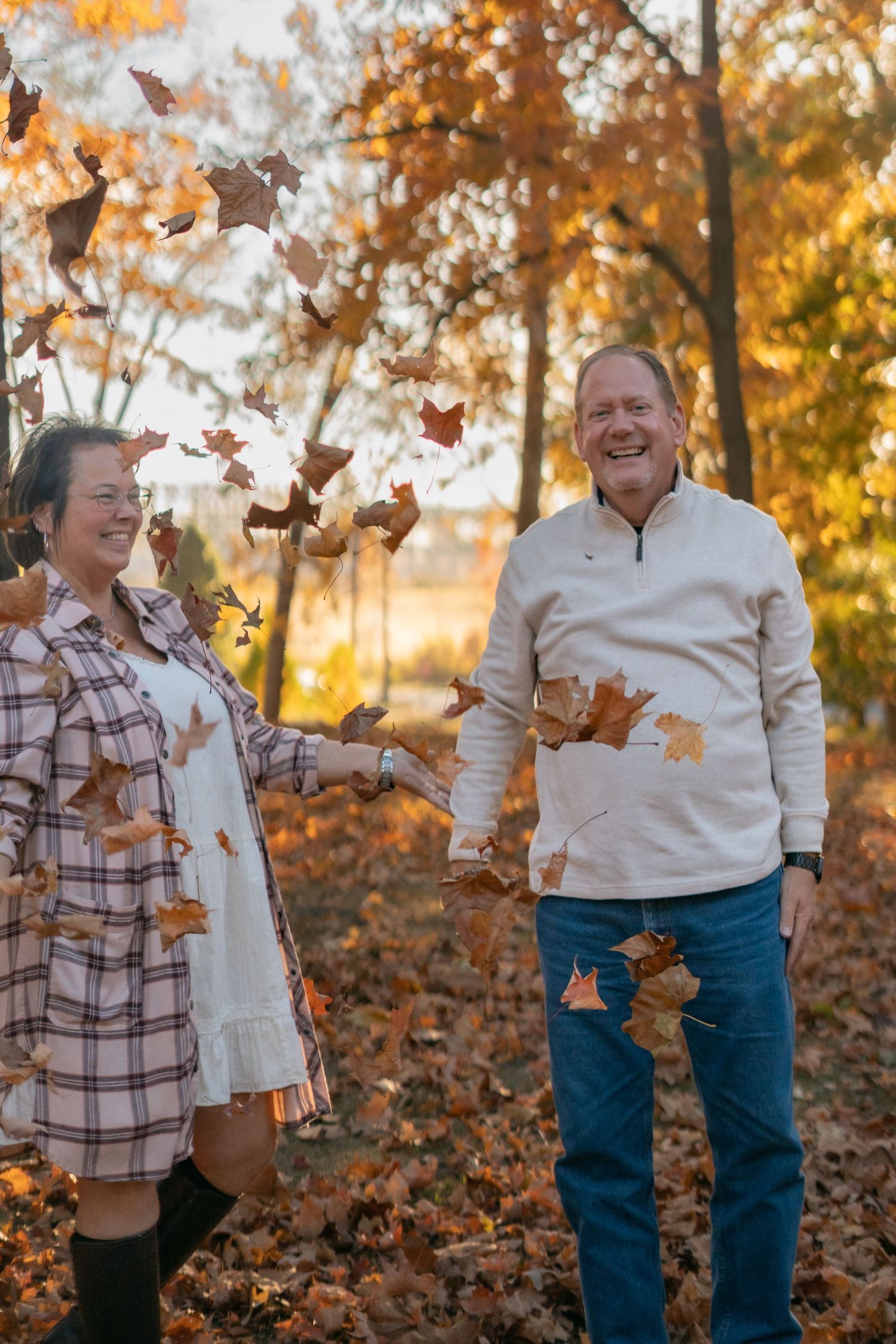 Two people joyfully toss fall leaves in the air during a golden sunset.
