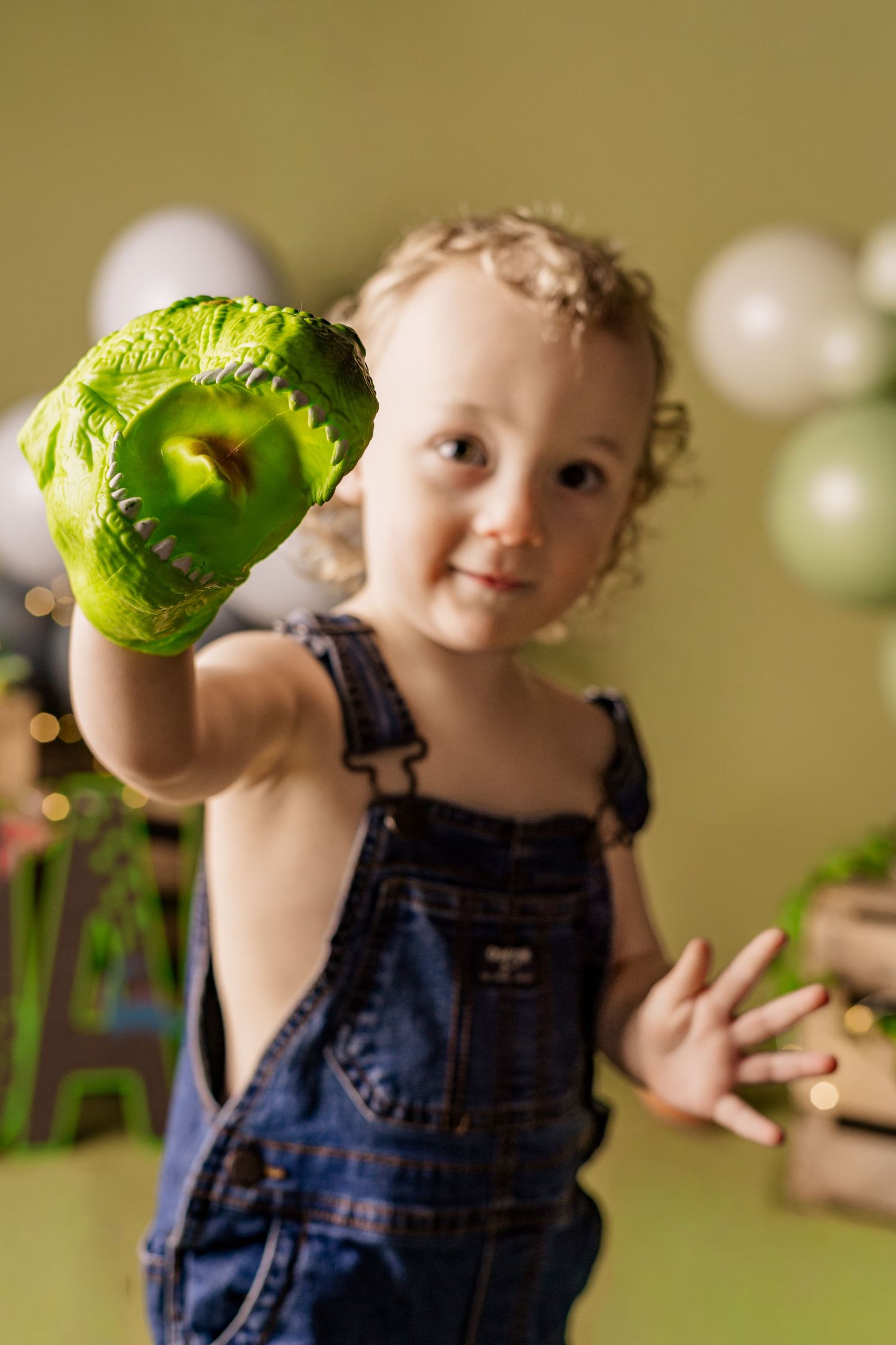 A young child in denim overalls shows off a bright green slime-covered hand against a blurred background.