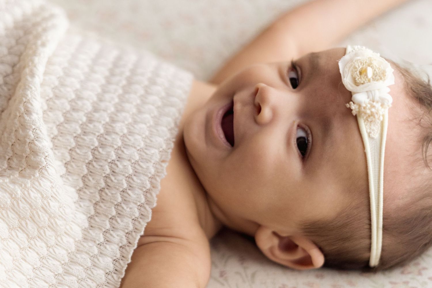 A small baby wearing a white headband with flower detail lies on textured white blanket while smiling.