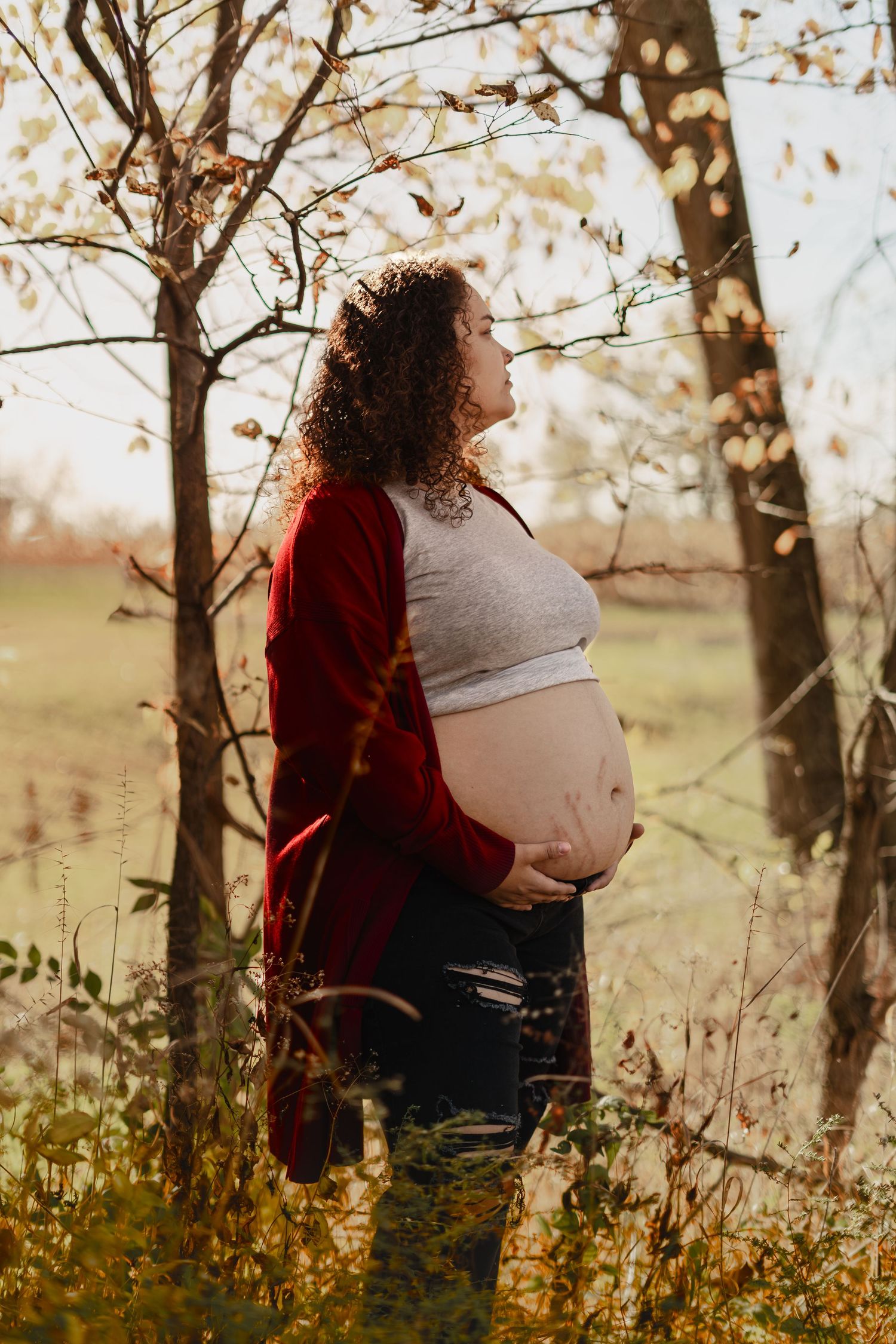 Side view maternity portrait with burgundy cardigan against golden sunset through tree branches.