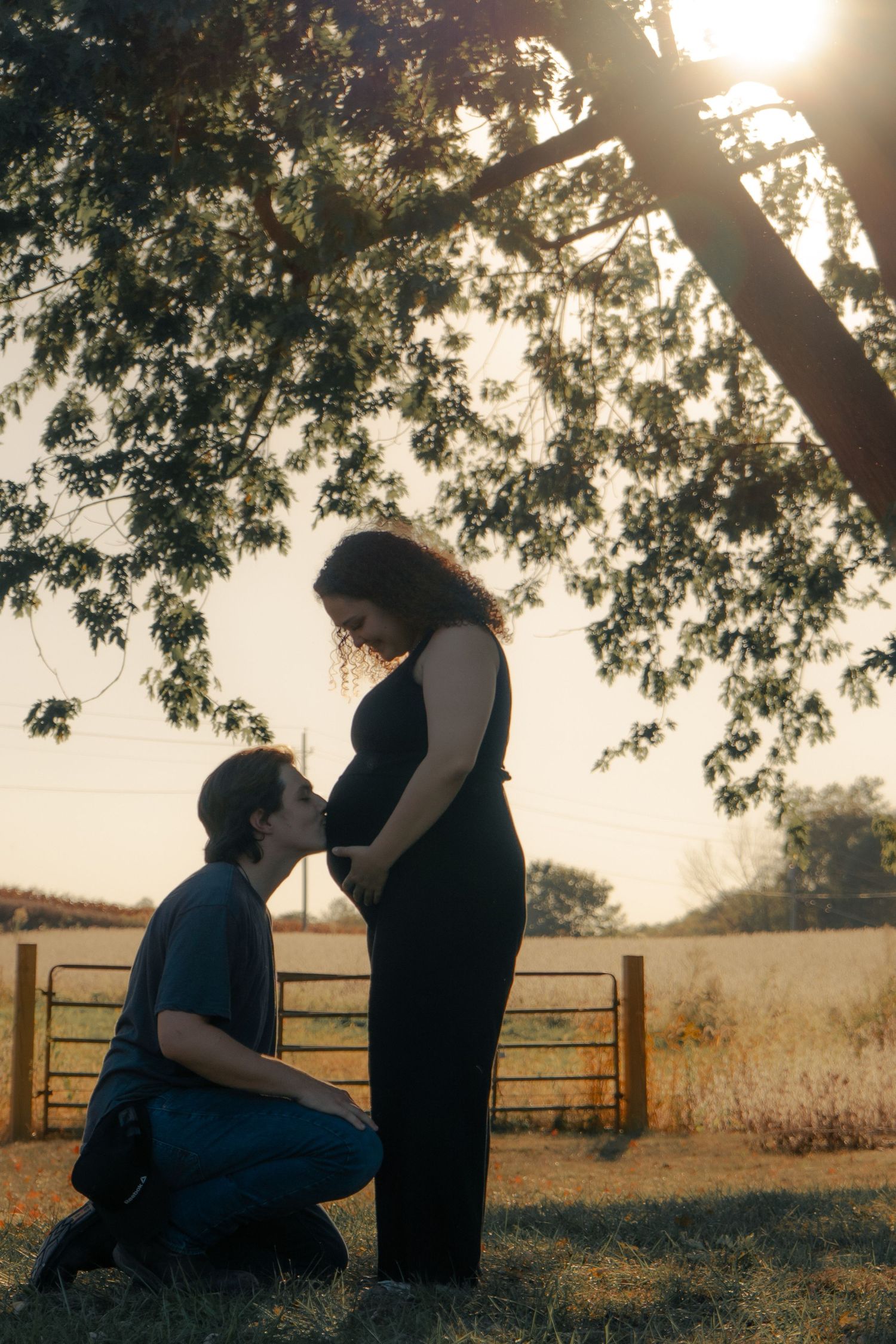 A silhouetted couple shares an intimate moment at sunset in a rural setting with trees and a fence in the background.
