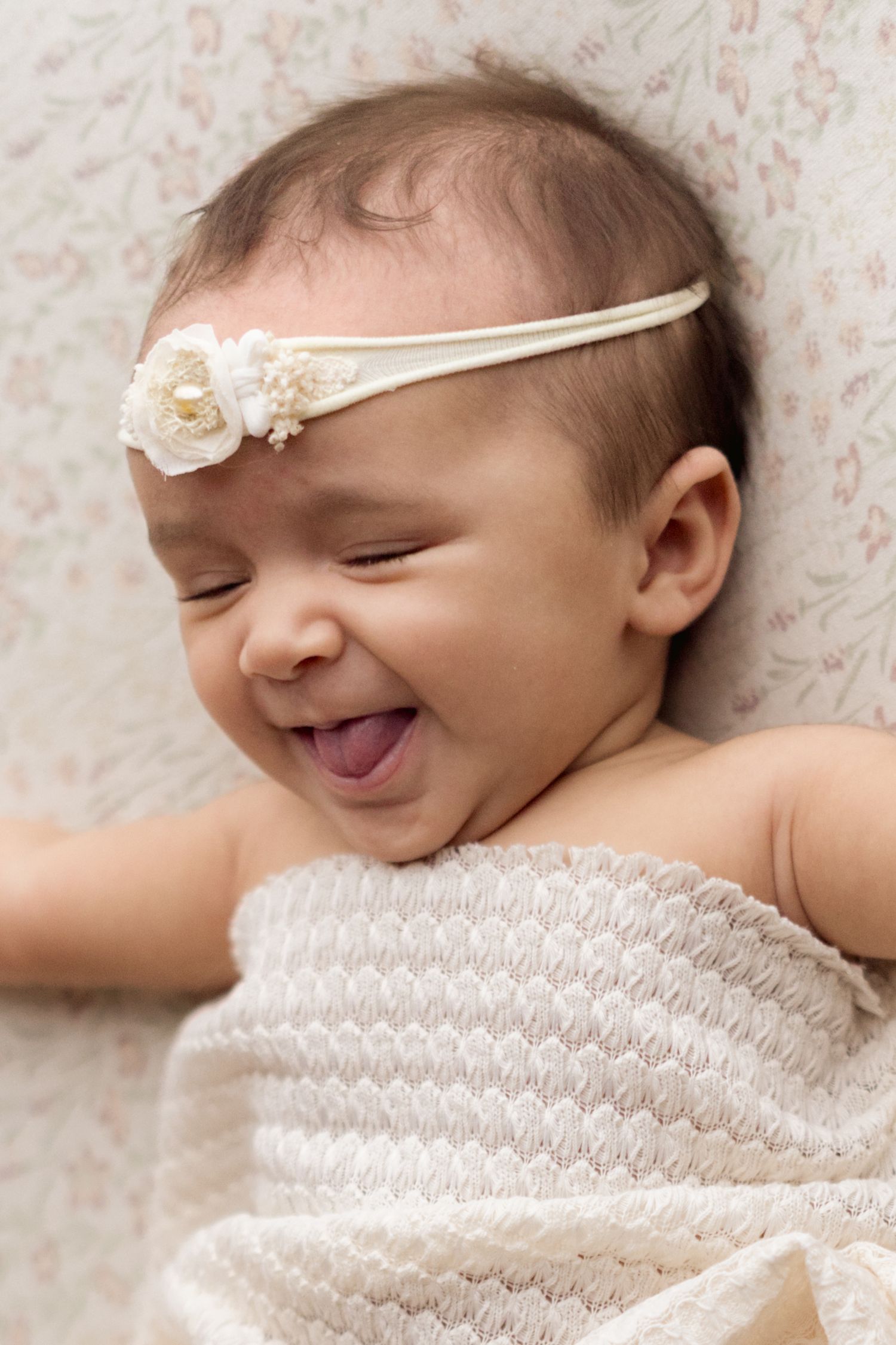 A sleeping newborn baby wrapped in delicate white lace smiles softly while resting on a floral background.