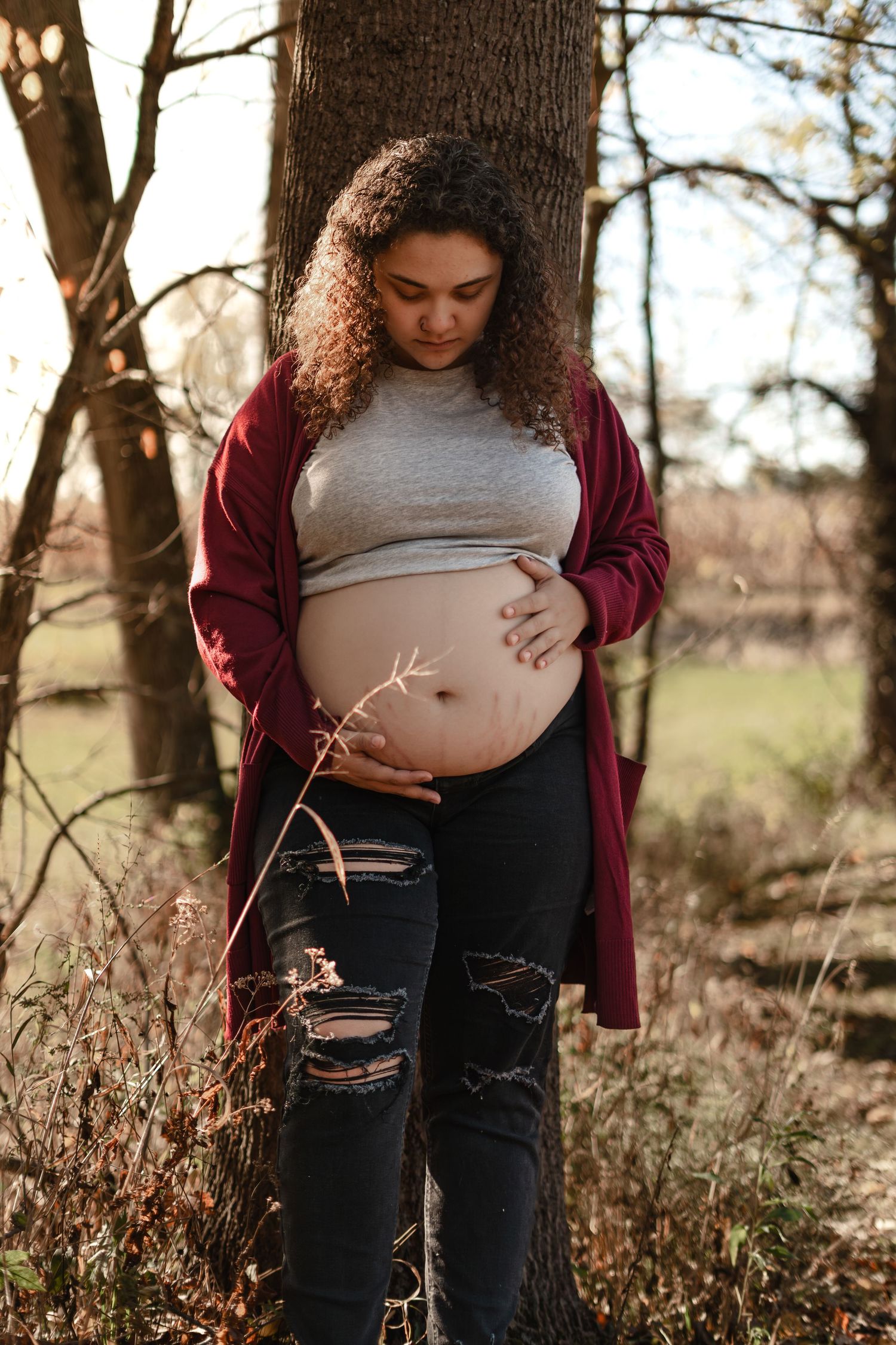 Intimate maternity portrait with burgundy cardigan and ripped jeans against natural backdrop.
