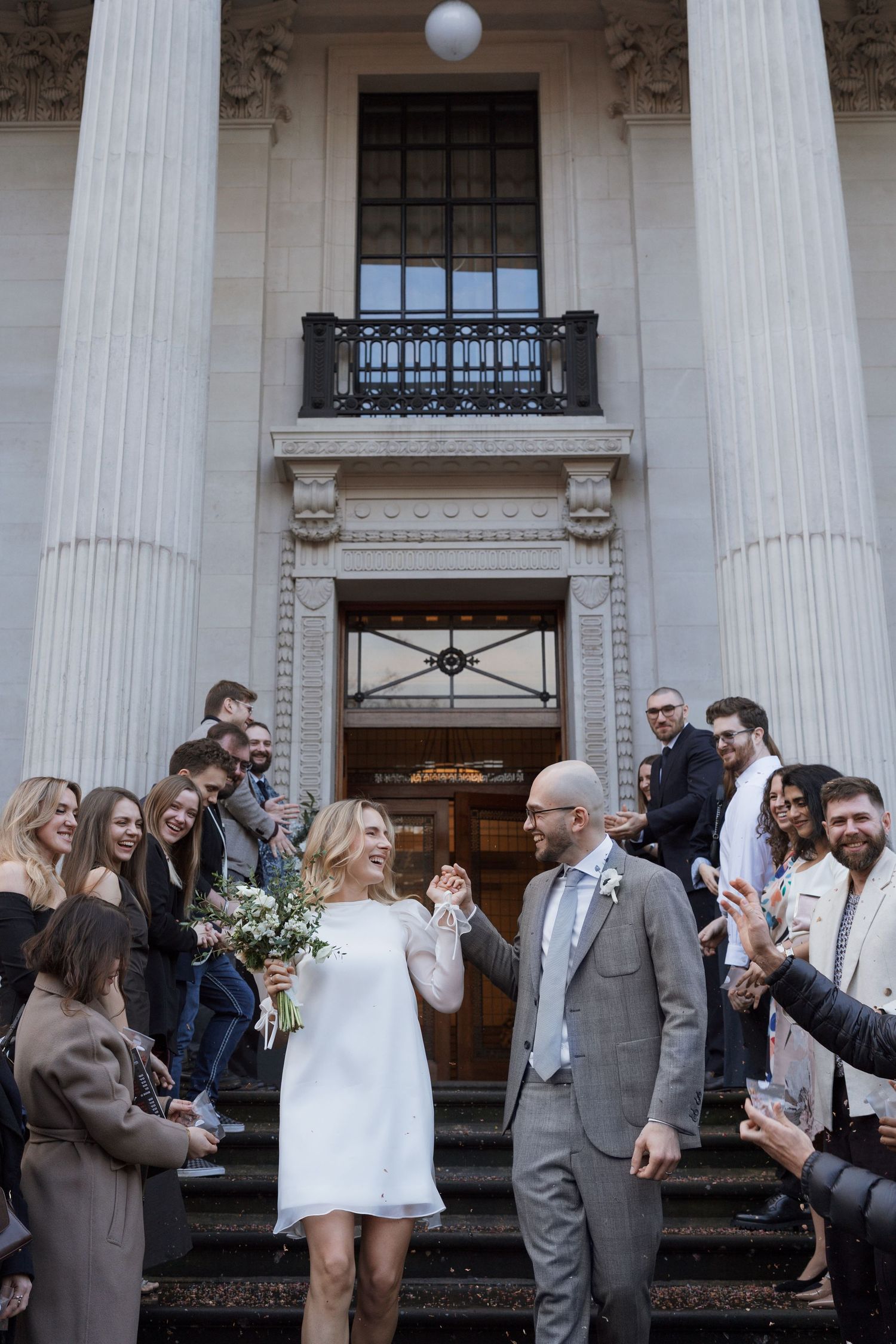 Wedding party celebrating outside a grand columned building with bubbles and confetti.