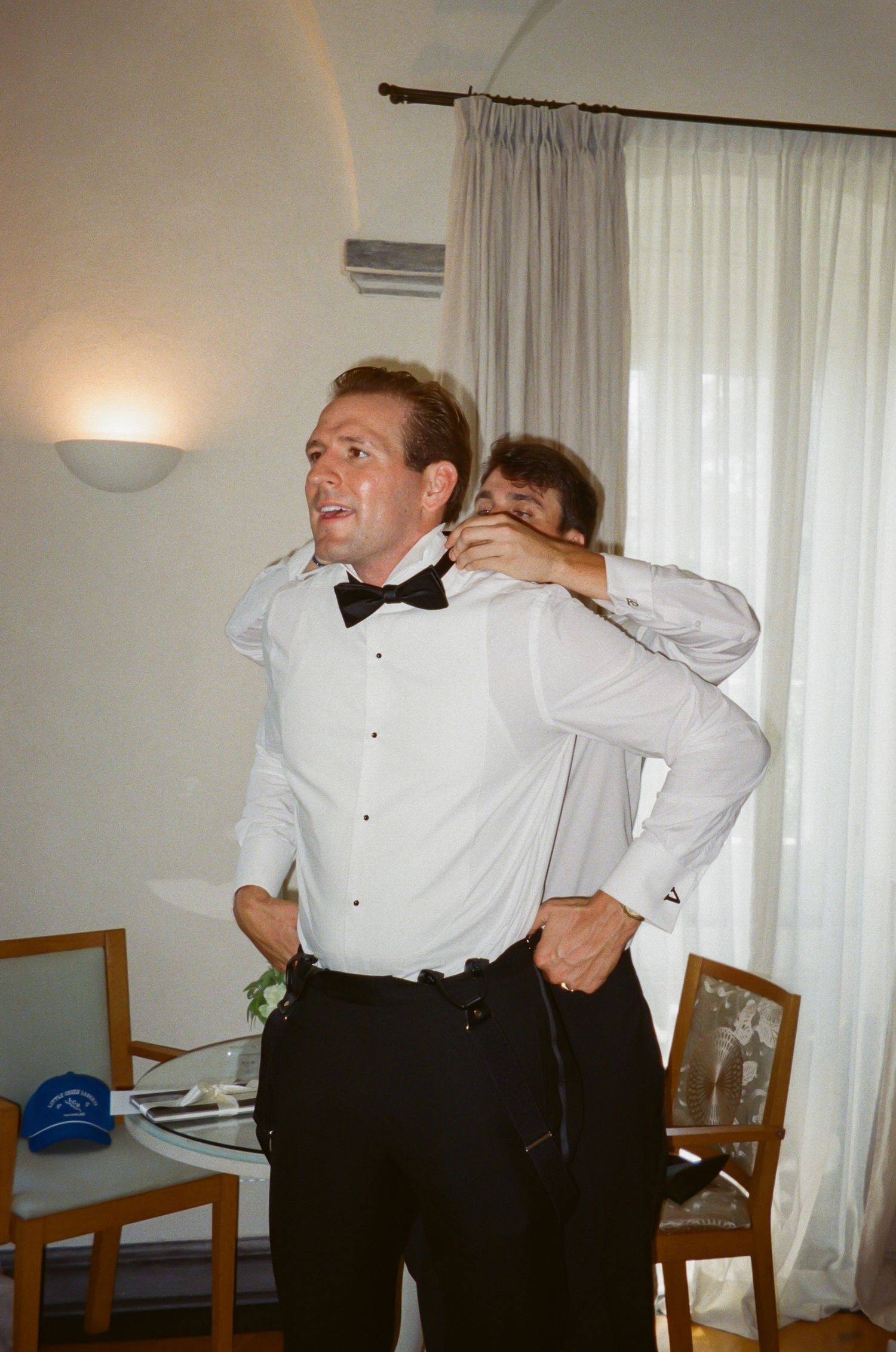 Two men in formal tuxedo attire preparing for a wedding, one adjusting the other's bow tie indoors.