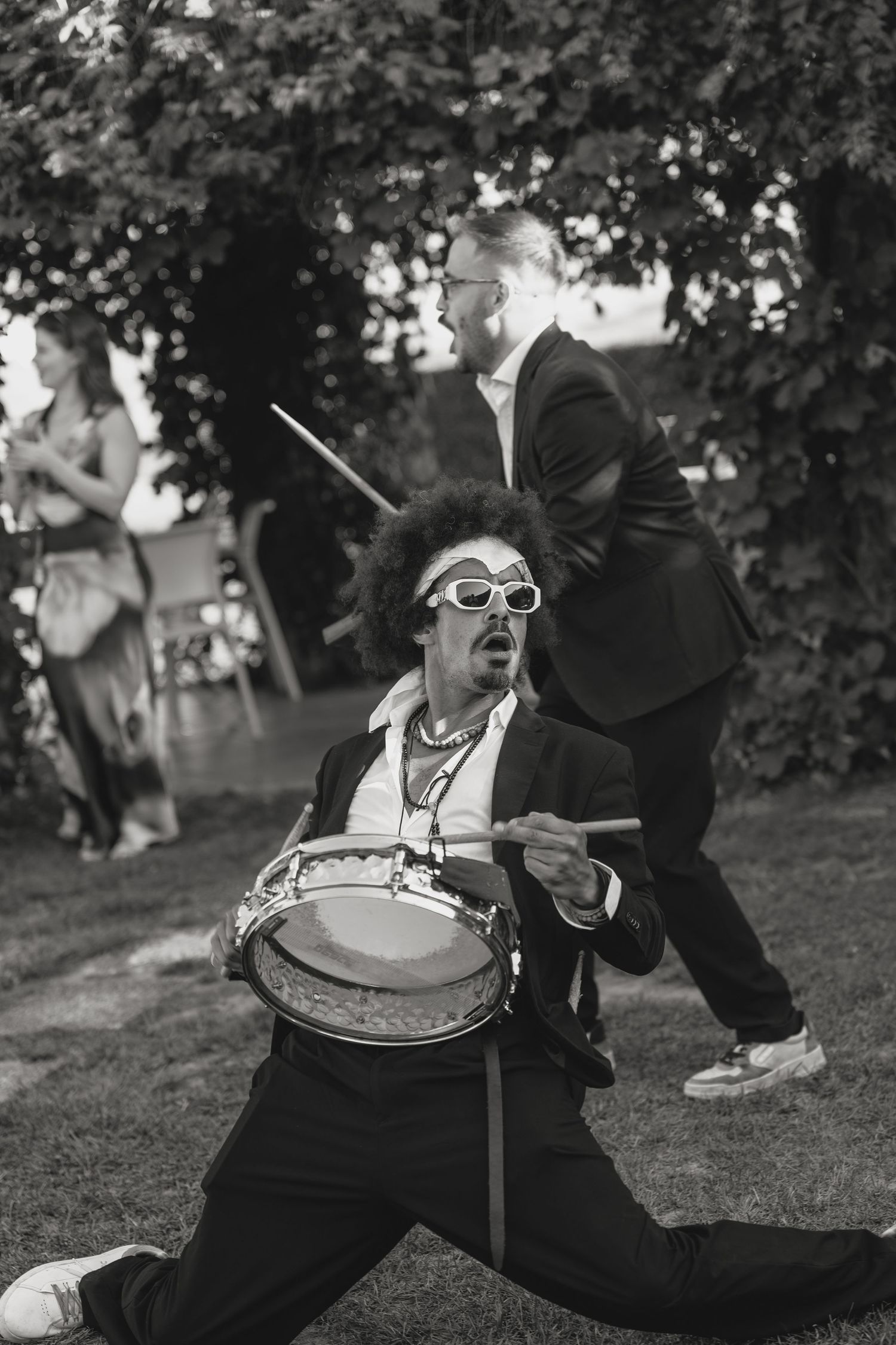 Black and white photo of an energetic drummer performing outdoors at a garden event, wearing sunglasses and an afro wig.