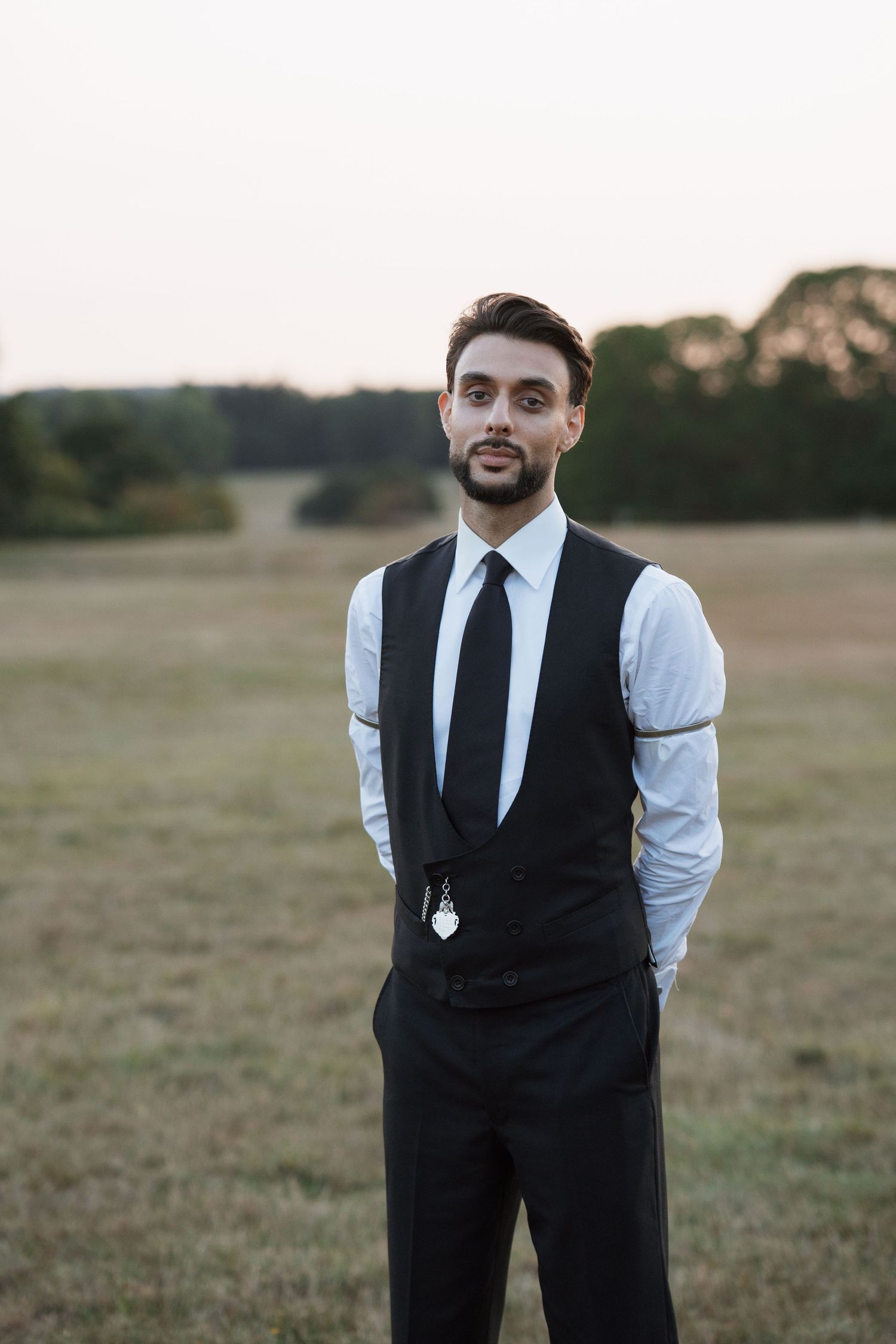 A person in formal attire with a black vest and tie stands in an open field at dusk.