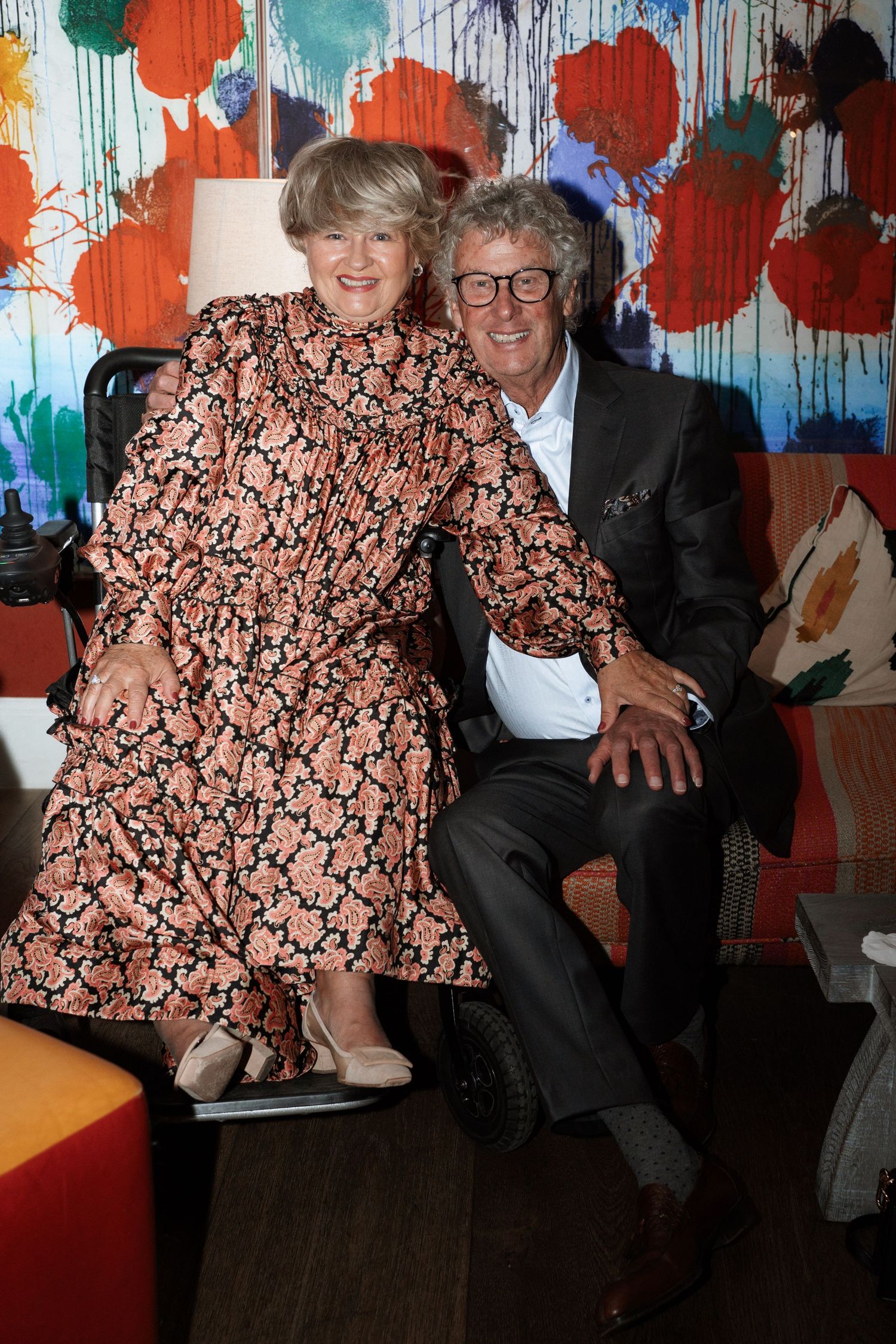 Two people sitting together against a colorful drip-painted wall background.
