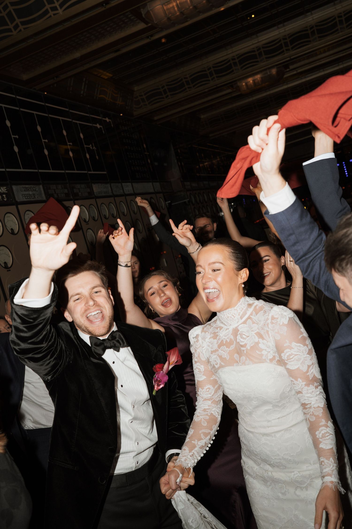 Joyful bride and groom dancing with guests at wedding reception, bride in lace gown, groom in black tuxedo.