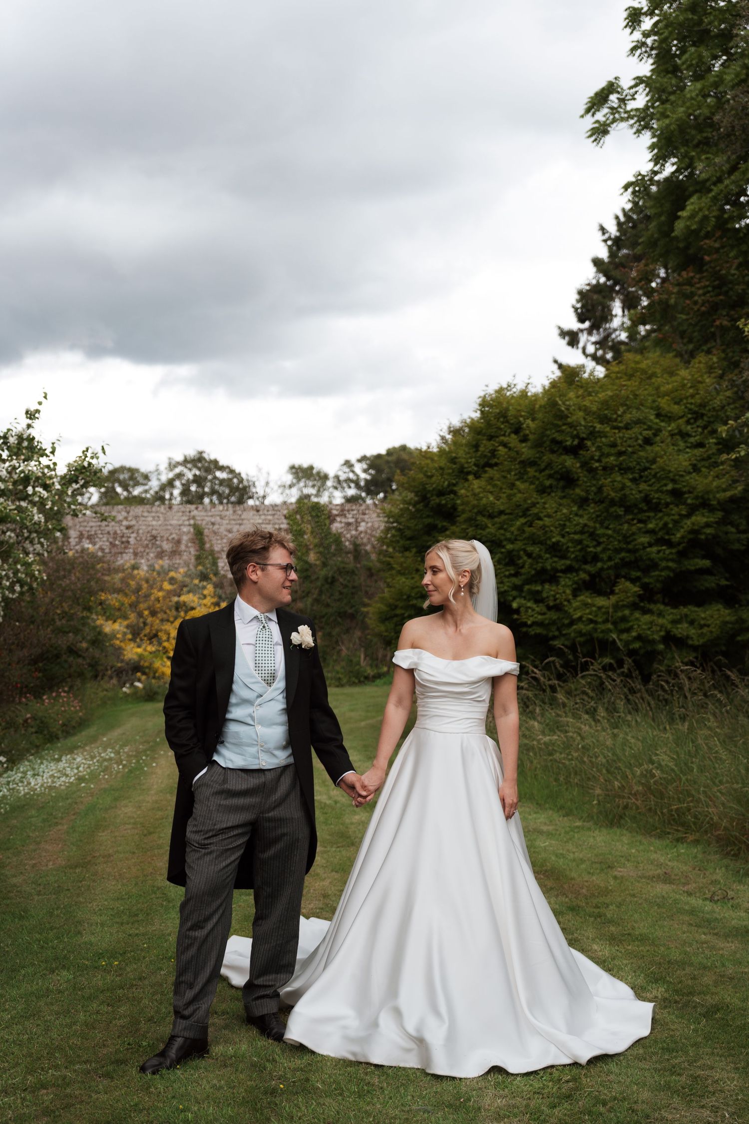 Bride and groom hold hands walking through lush garden grounds, bride in off-shoulder white gown, groom in morning suit.