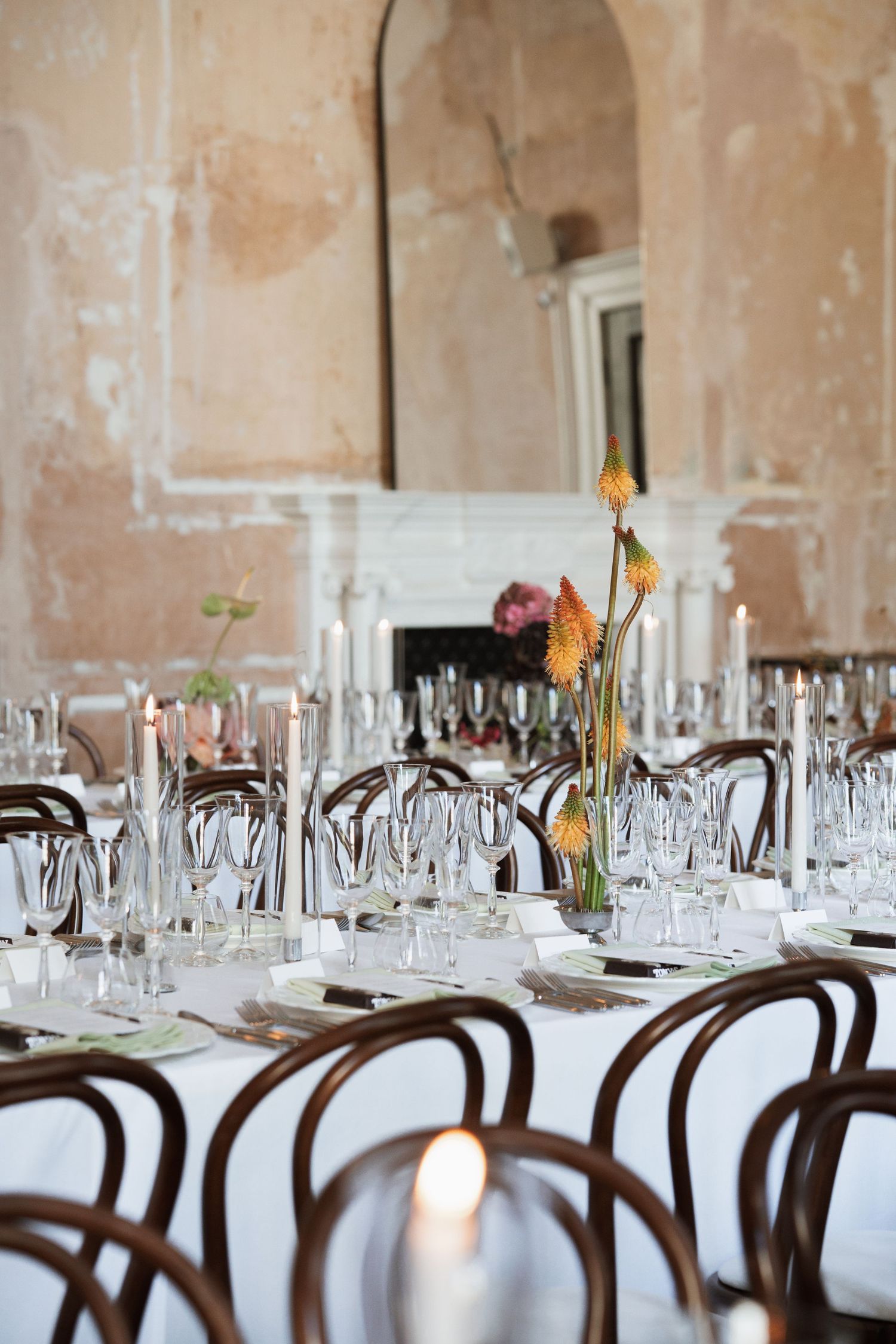 Elegant wedding reception table with candles, glassware and orange flowers in a rustic venue with a white fireplace and bentwood chairs.