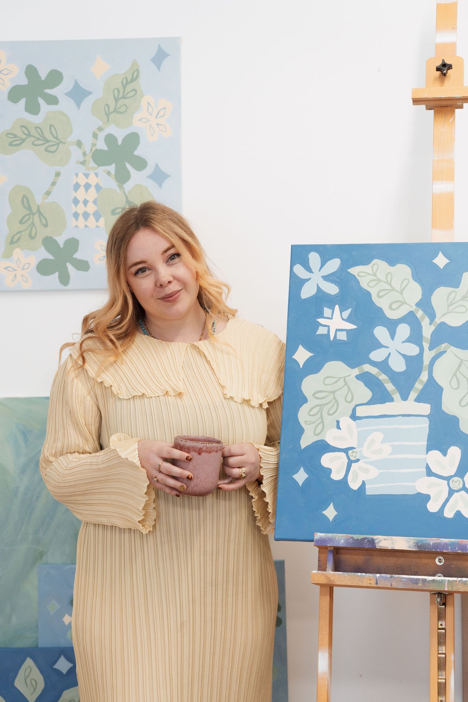A blonde woman in a yellow dress holds a mug beside blue floral paintings on easels in a bright art studio.