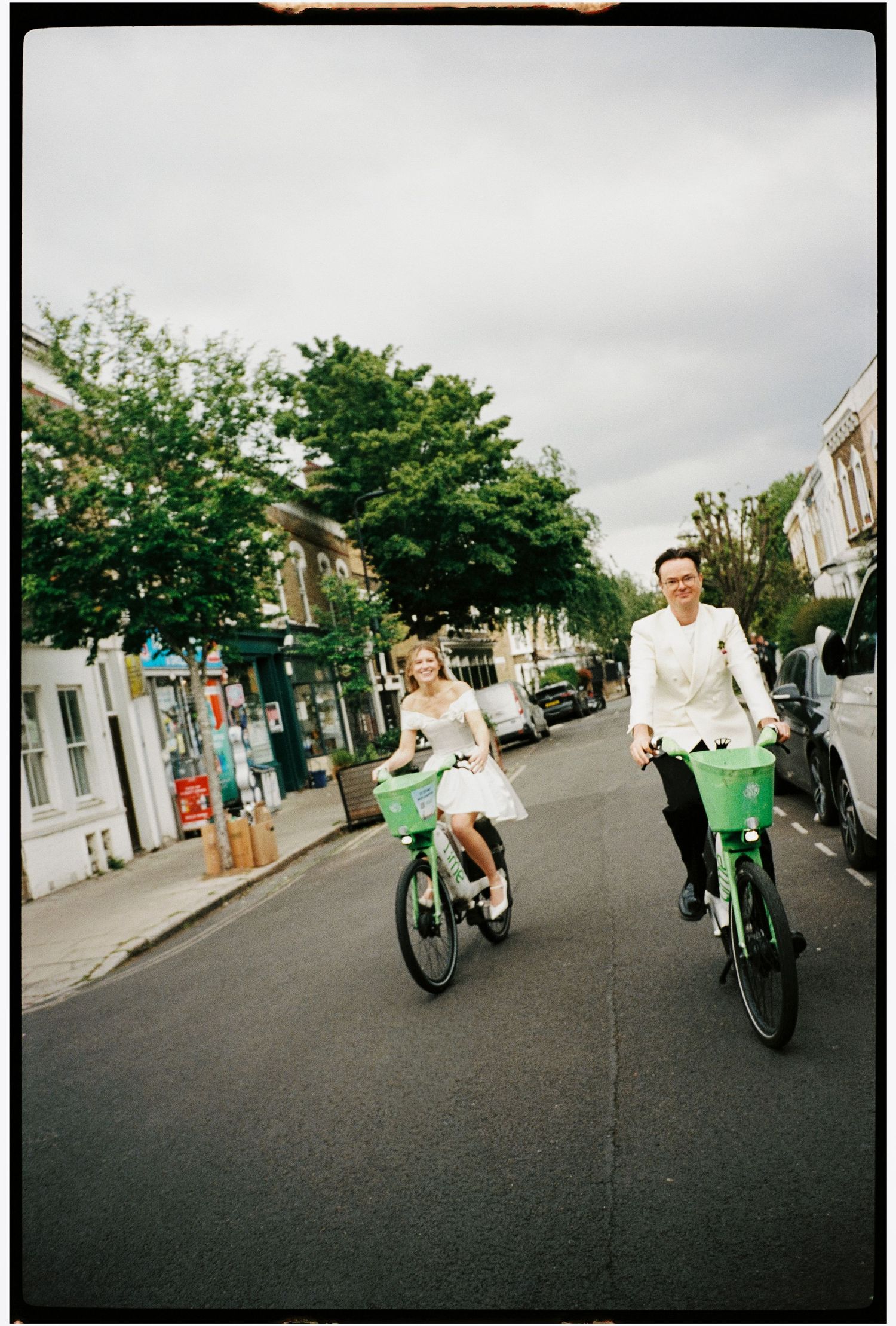 A bride and groom ride green bikes down a tree-lined street in wedding attire on a cloudy day.