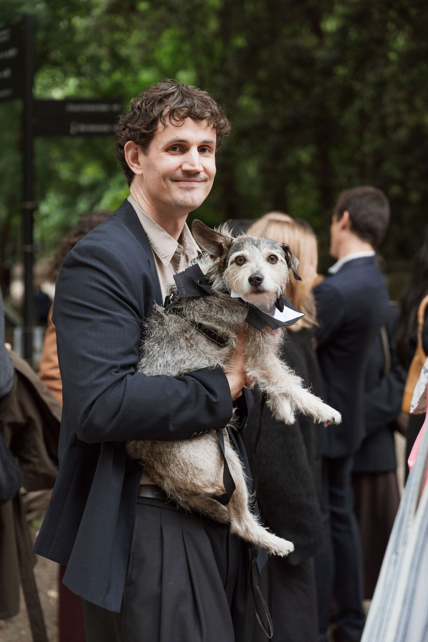 Smiling man in dark suit holding a small scruffy dog dressed in a bow tie at an outdoor event.