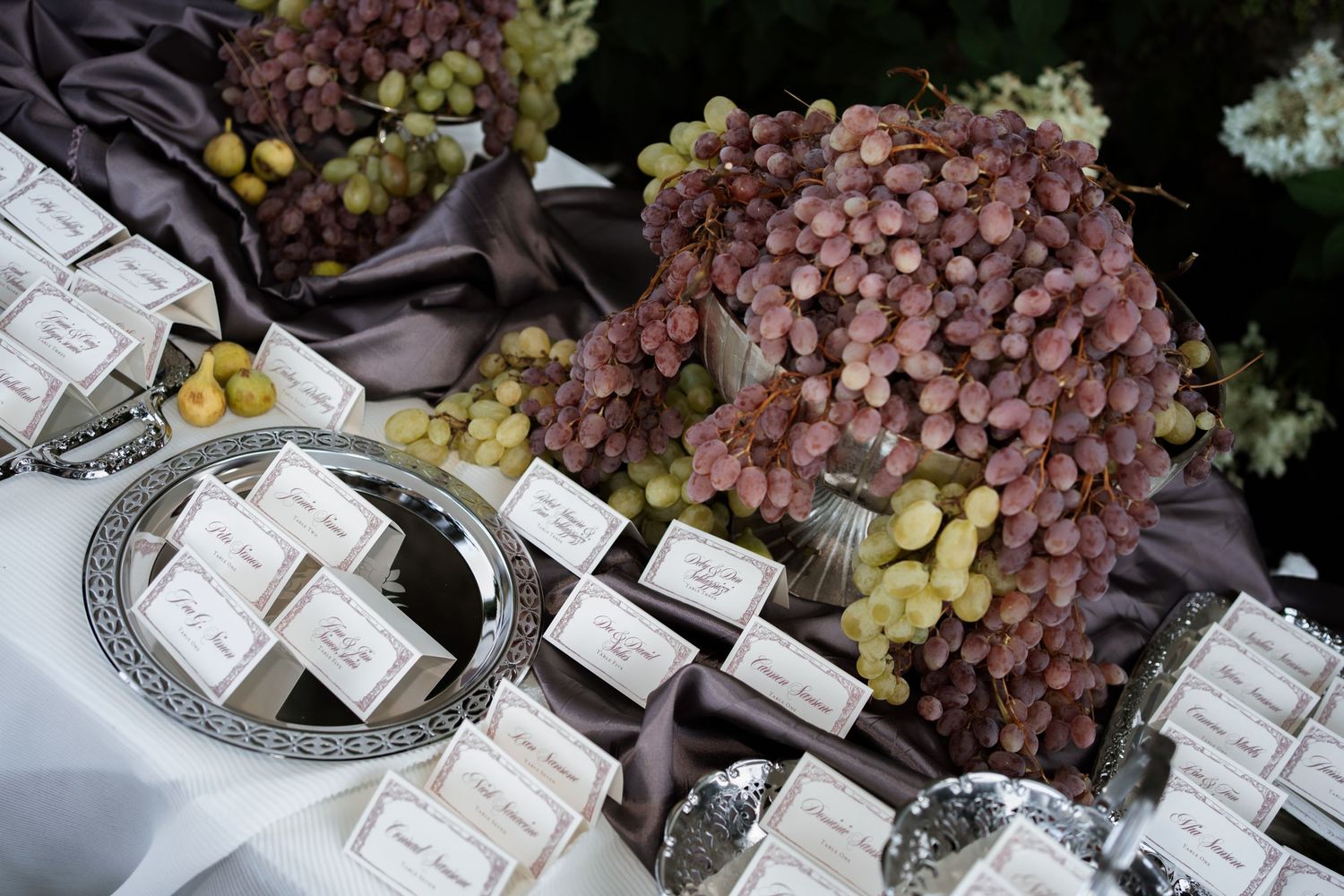 Elegant wedding escort cards displayed on silver trays beside decorative grape clusters on a white tablecloth.