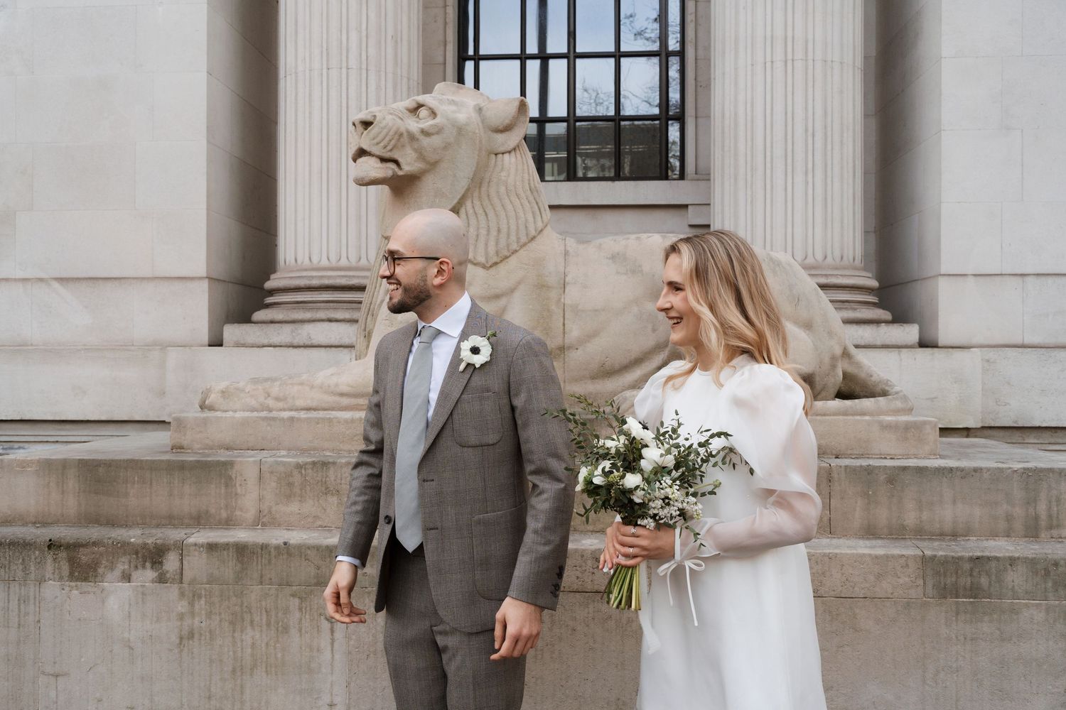 A wedding couple poses in front of a stone building with classical architecture and a lion statue.