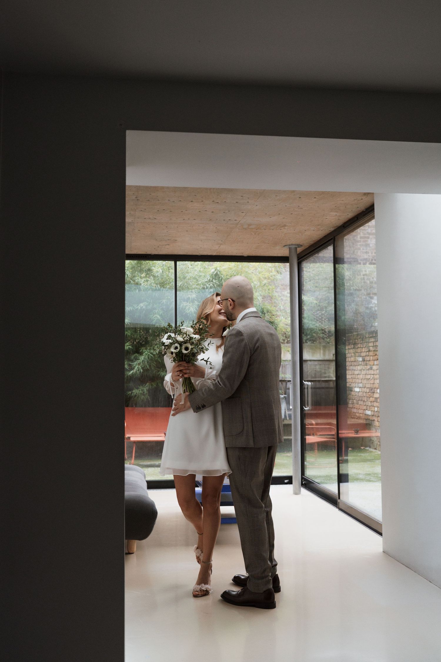 Wedding couple embracing in a modern room with large windows and white walls.