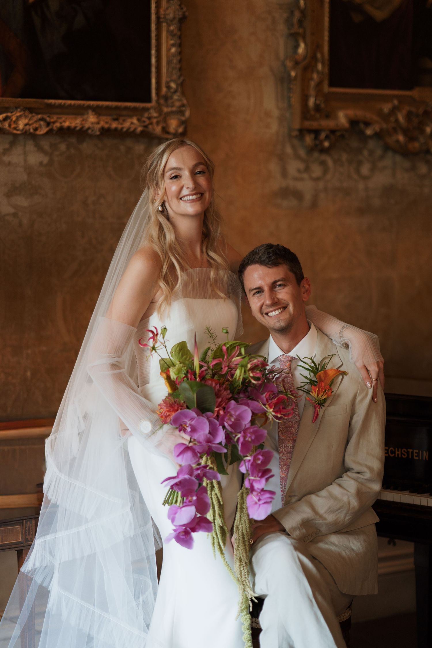 Wedding couple poses with vibrant pink and purple floral bouquet in an ornate room.