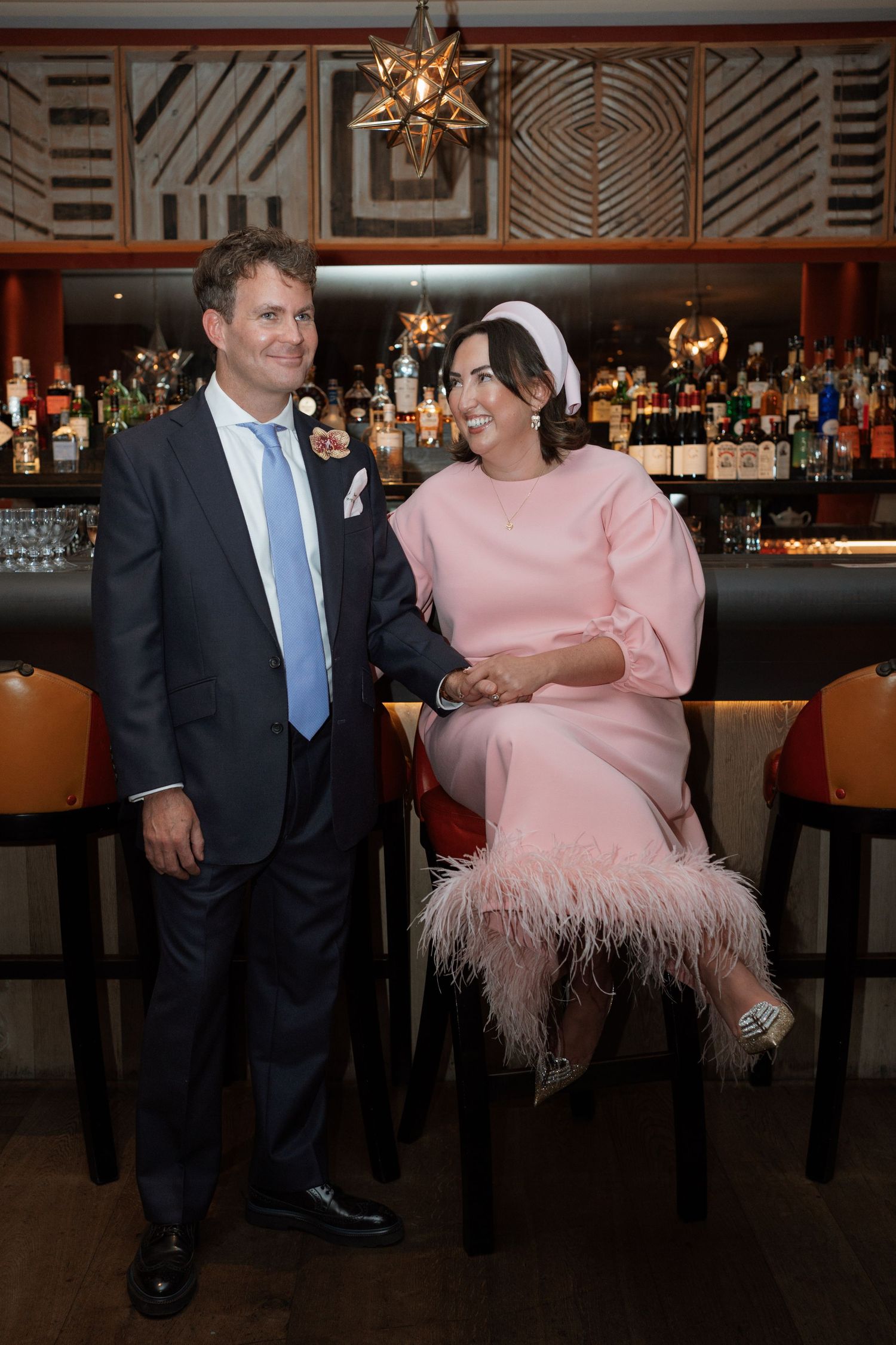 Couple in formal attire posing at a bar with geometric wall patterns.