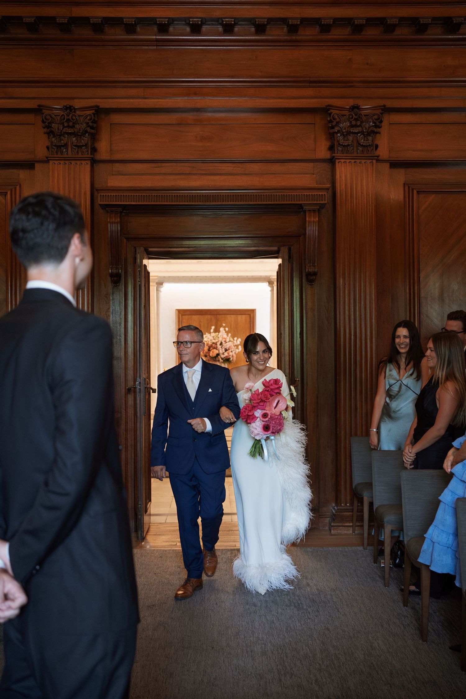 Wedding couple walks through elegant wood-paneled hallway with guests looking on.