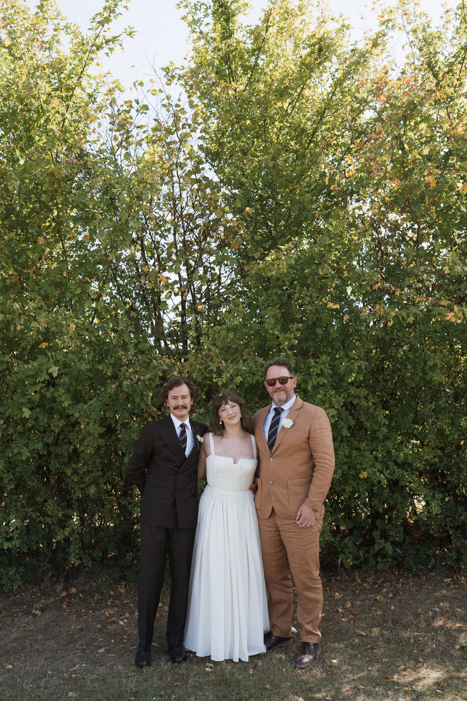 Wedding group portrait taken outdoors with leafy green trees as backdrop.