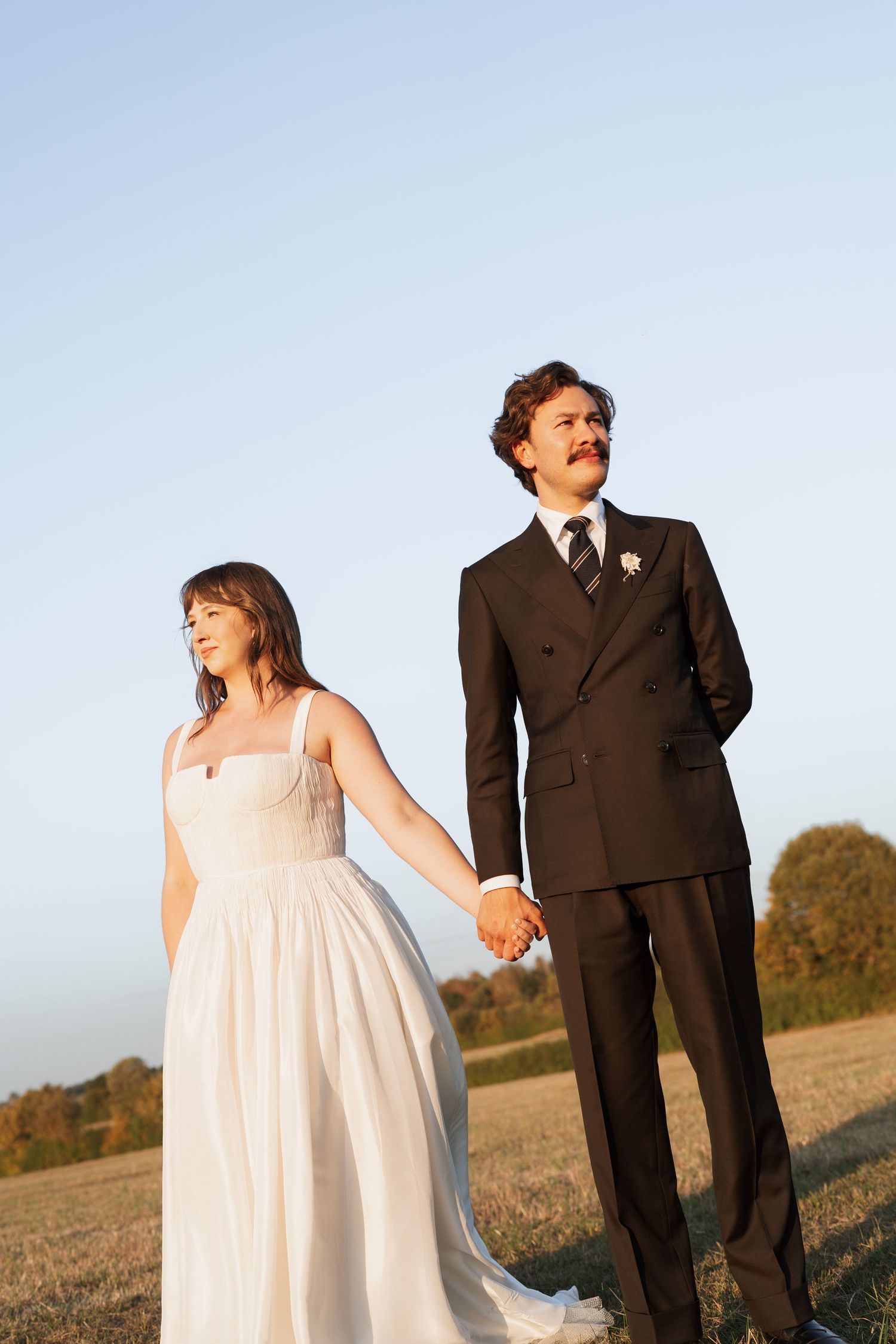 Wedding couple posing in a sunlit field with autumn trees in the background.