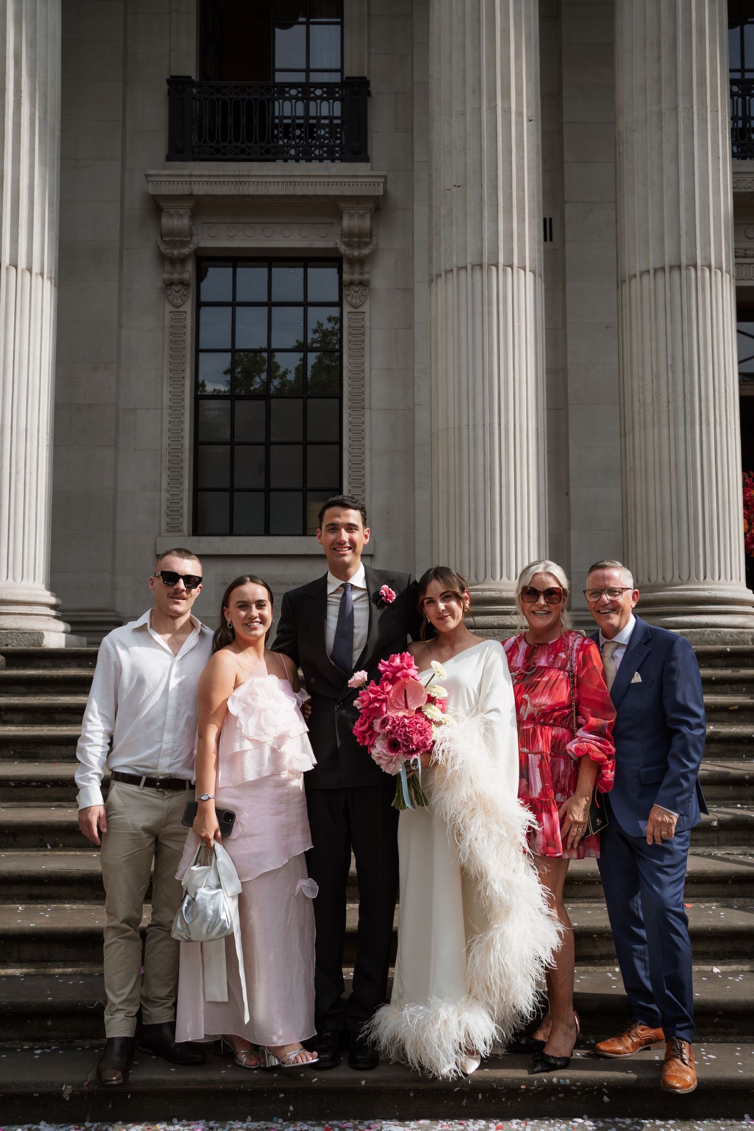 Wedding group poses for formal photo in front of classical columned building.