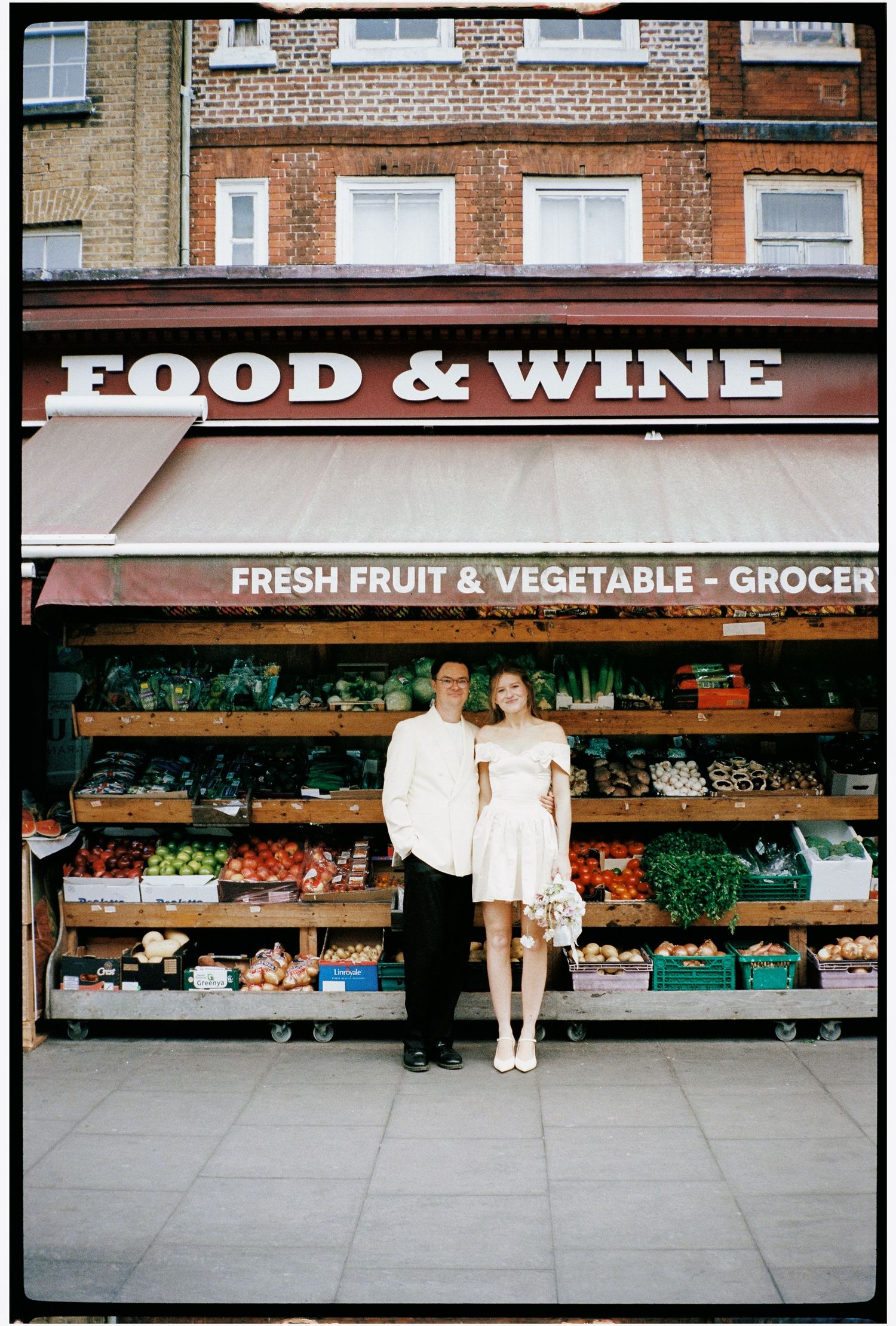 Couple in wedding attire posing in front of a Food & Wine fresh fruit and vegetable grocer shop.
