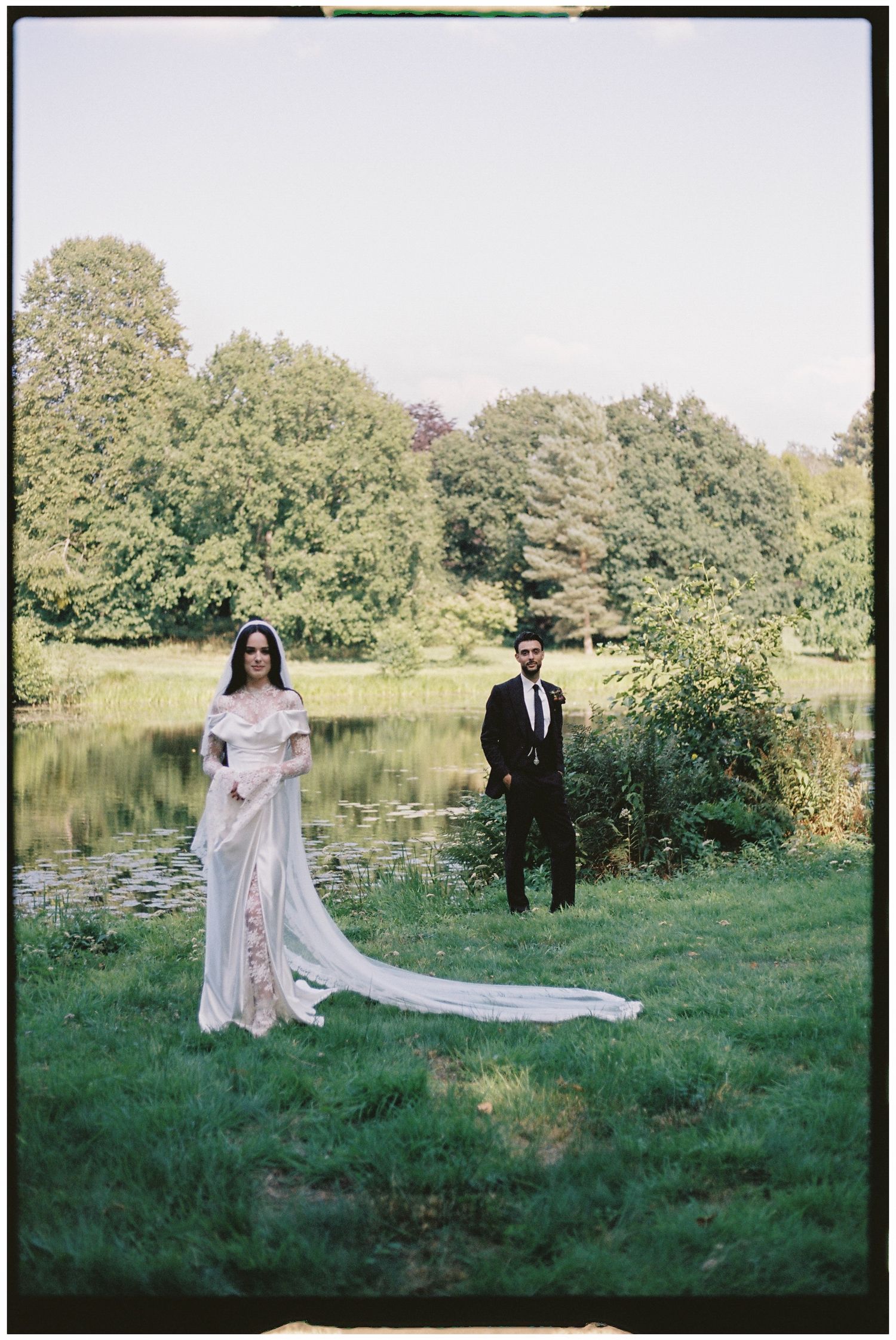 Wedding couple in formal attire posing by scenic pond with long flowing veil.