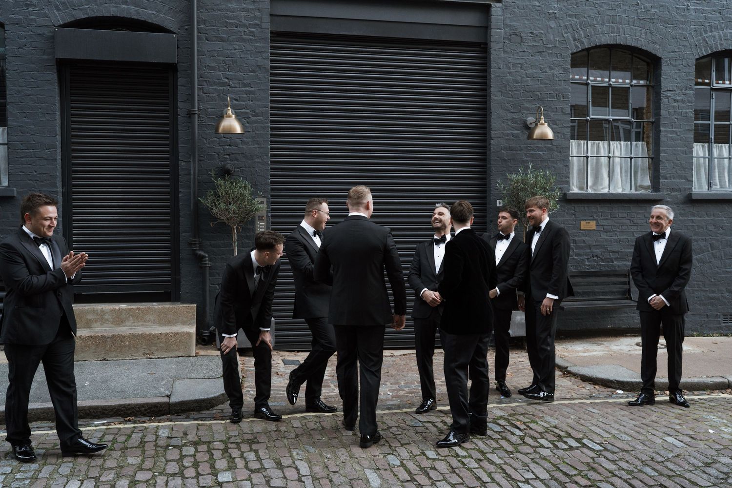 Groomsmen in black tuxedos pose together on a cobblestone street.