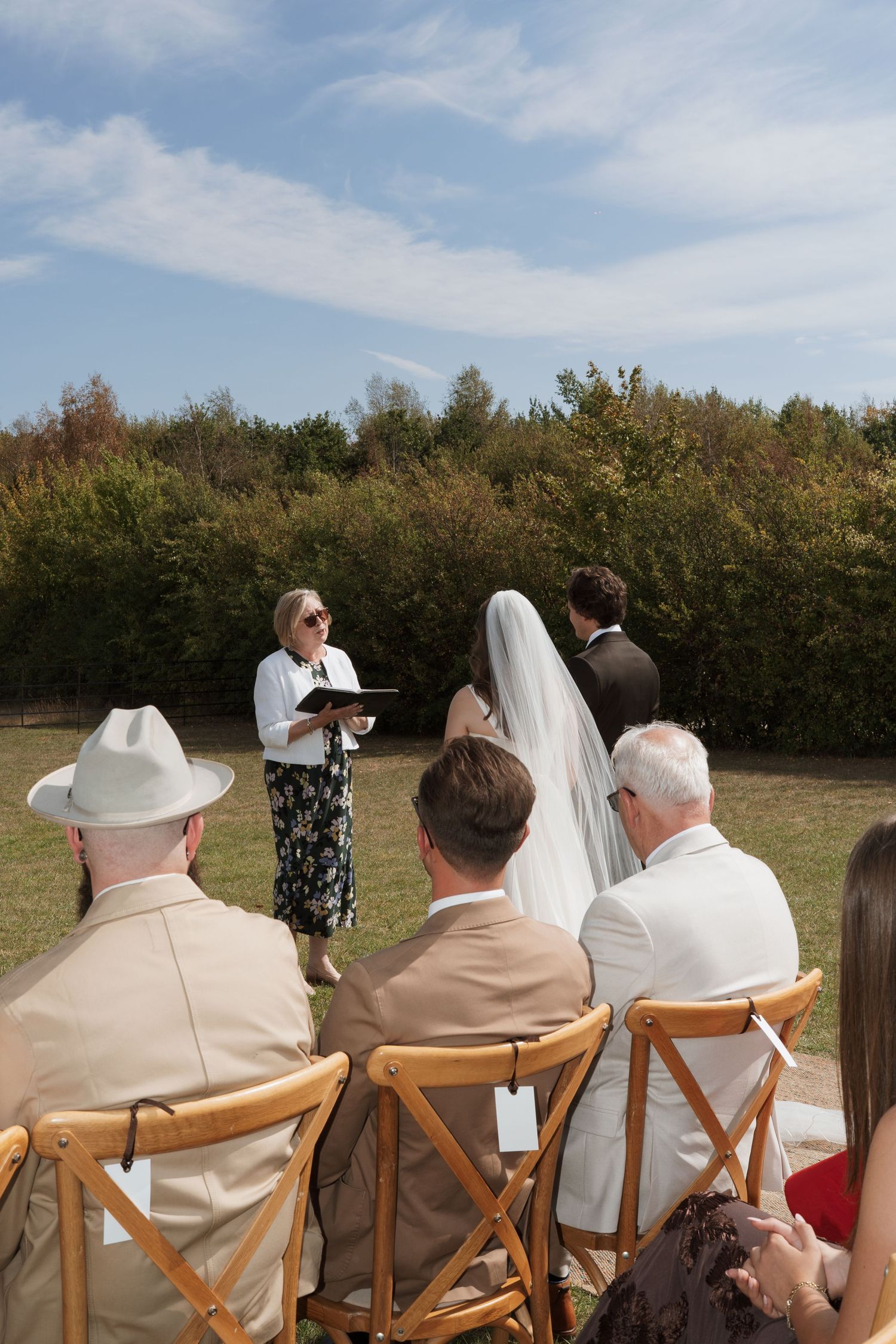 Wedding guests seated in wooden chairs watch outdoor ceremony against hedgerow backdrop.