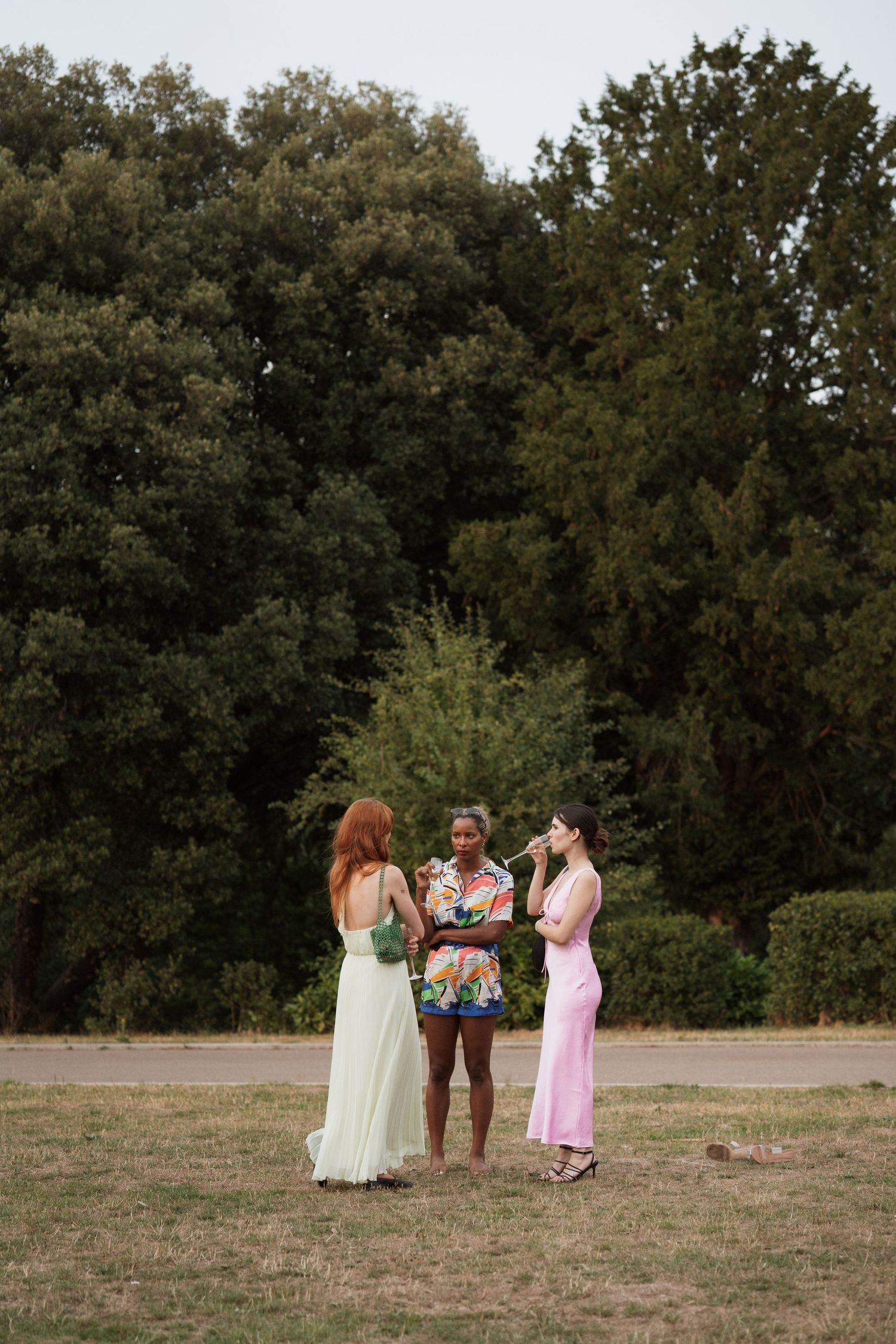 Wedding guests in pastel dresses gathering on a grassy field with trees in the background.