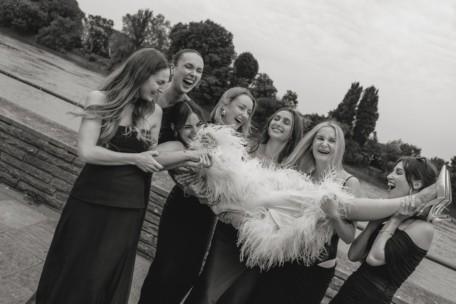 A joyful wedding party laughs together outdoors on a cloudy day.