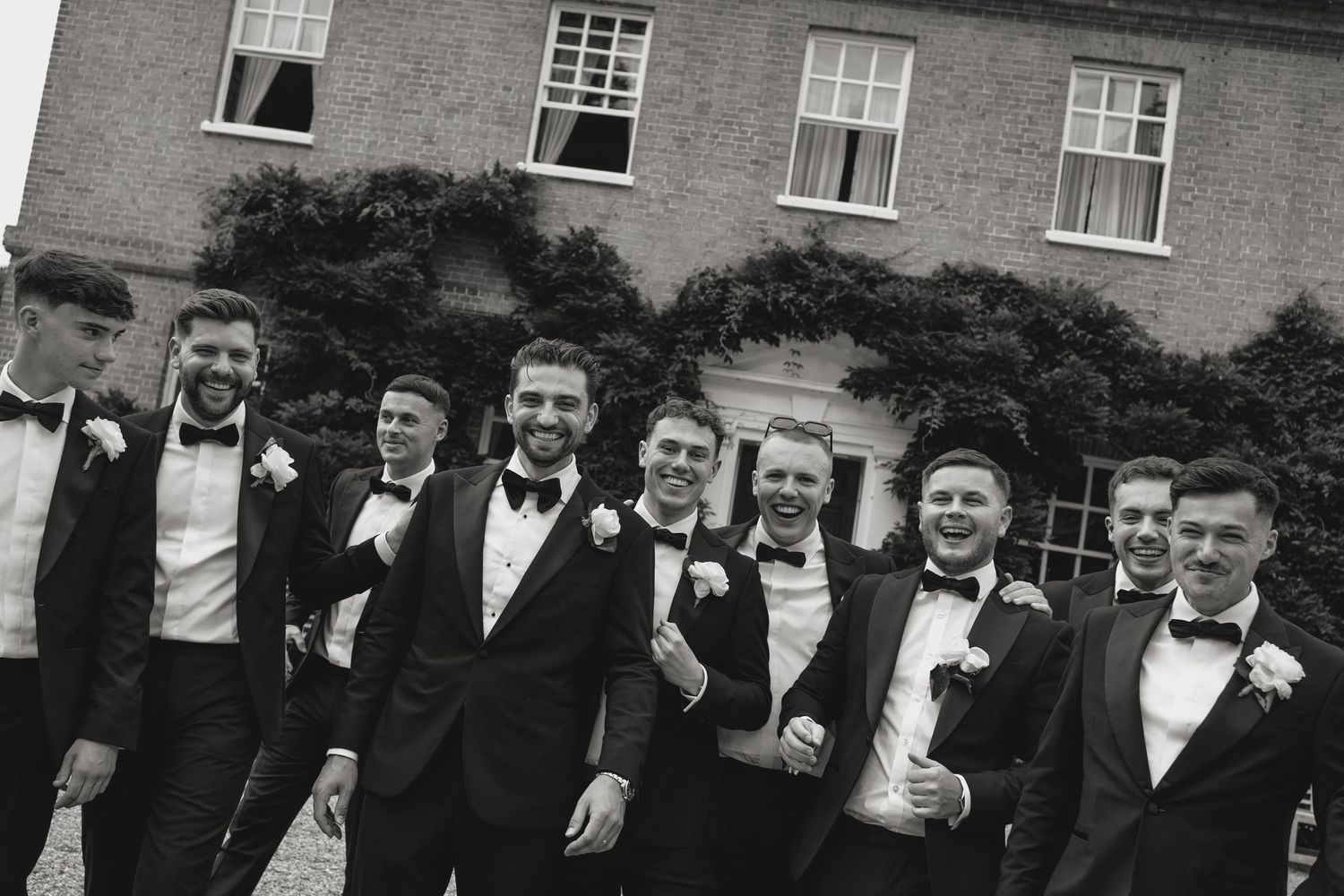 A black and white group photo of groomsmen in tuxedos posing in front of a brick building.