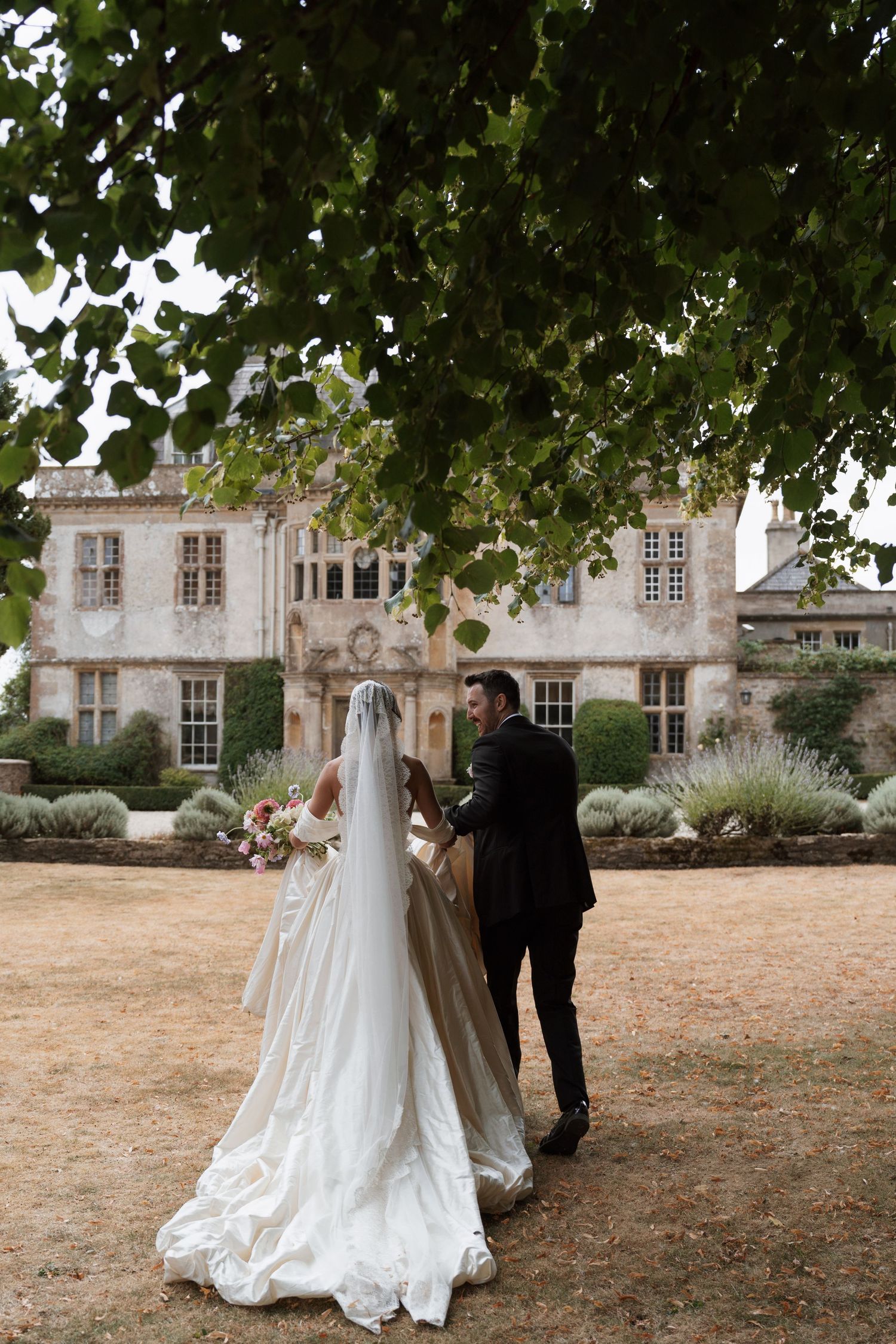 Bride in a long white veil and satin gown walks with a groom in a dark suit toward a historic stone manor on a sunlit lawn.