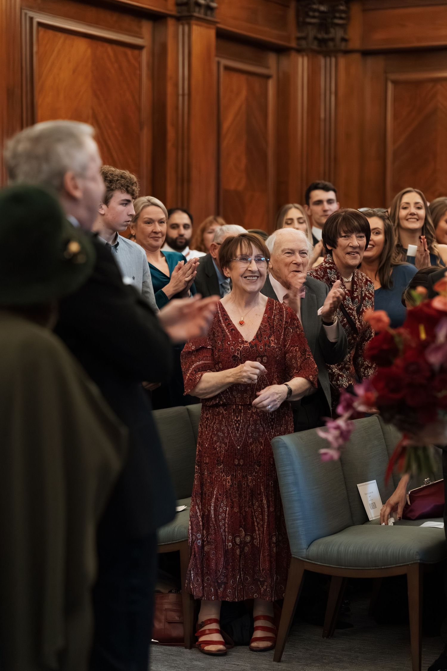 Wedding guests standing and applauding in a wood-paneled ceremony room.