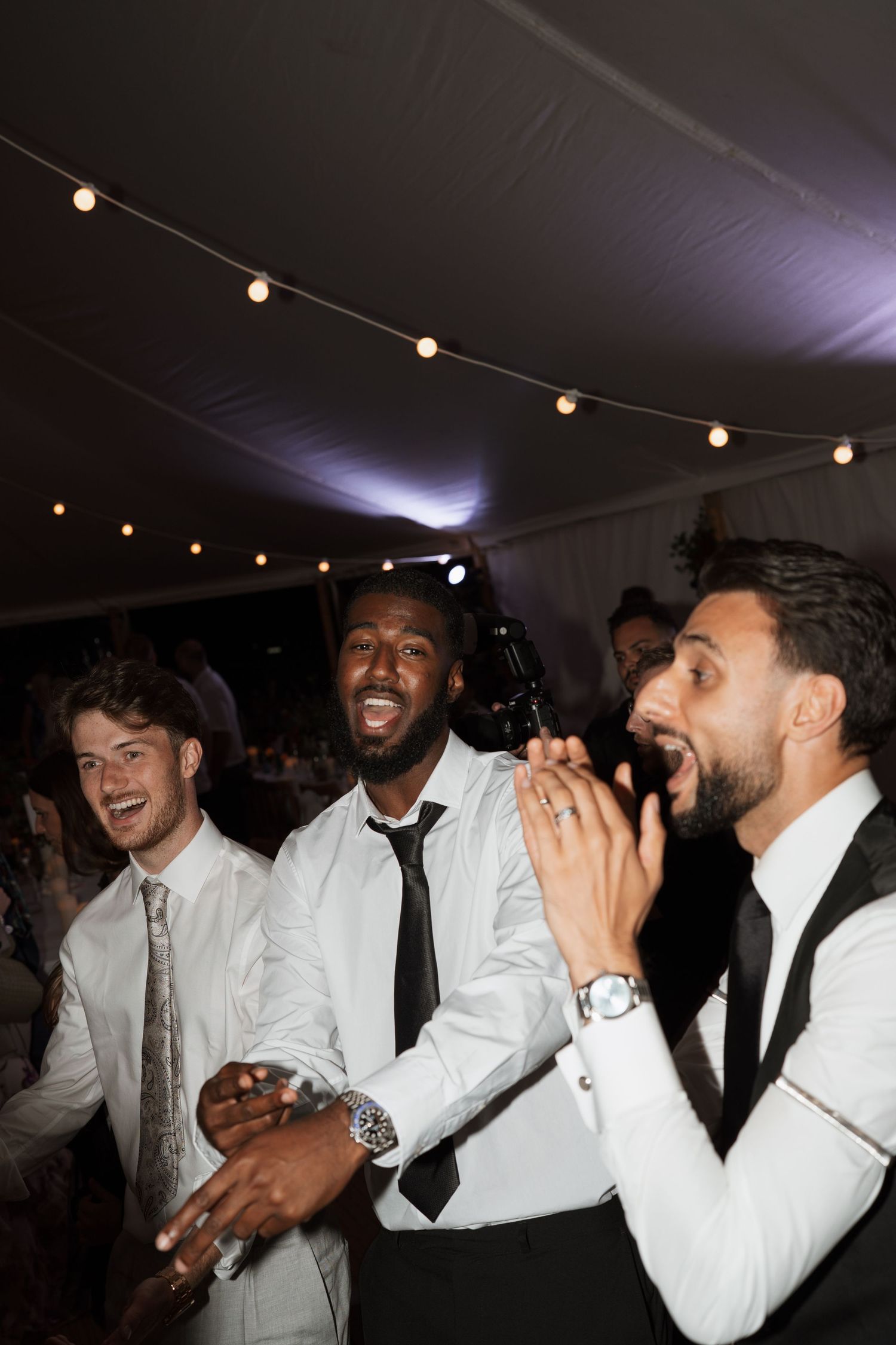 Wedding guests in white shirts and ties celebrate on the dance floor under string lights.