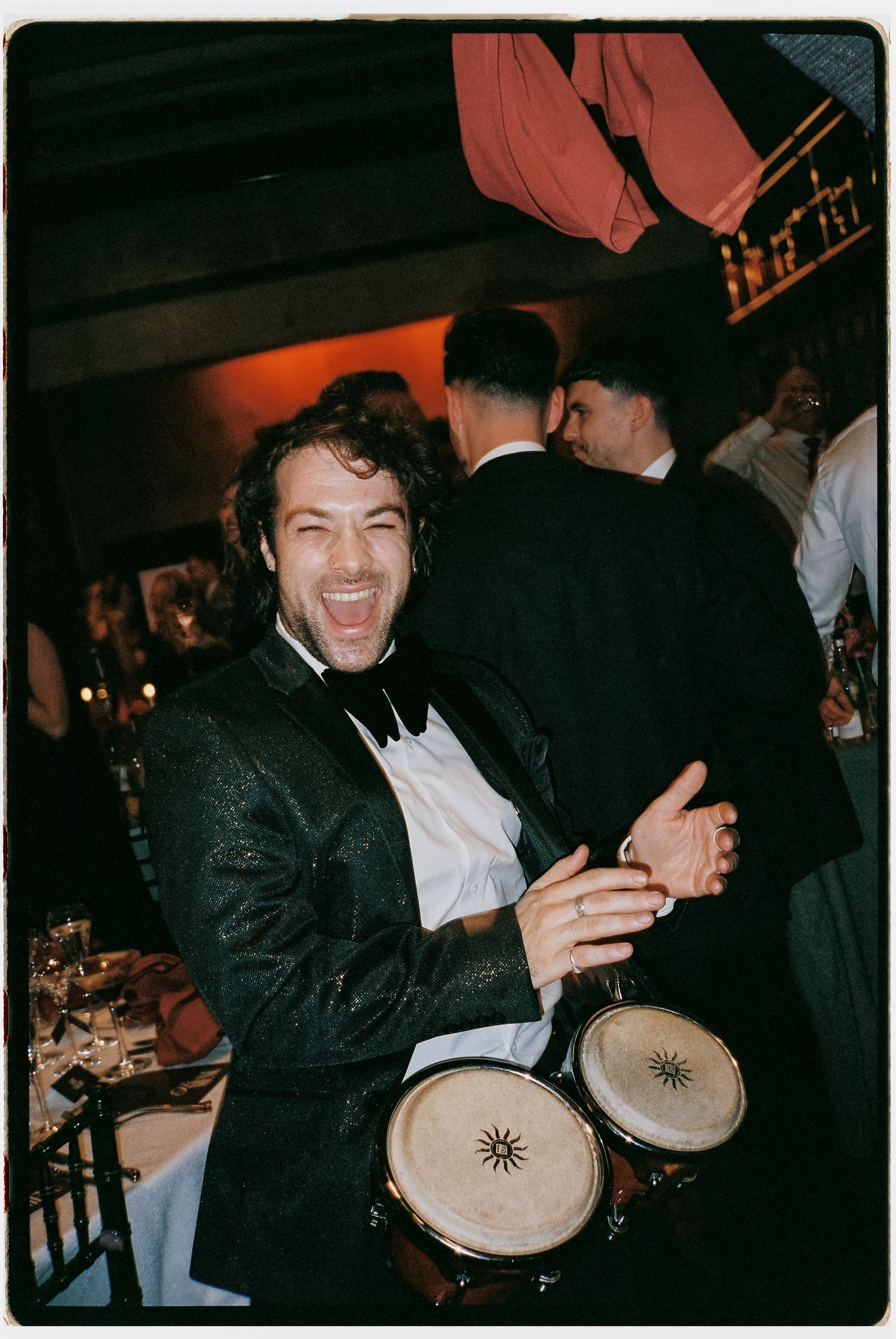 Enthusiastic guest in tuxedo playing bongo drums at formal celebration.