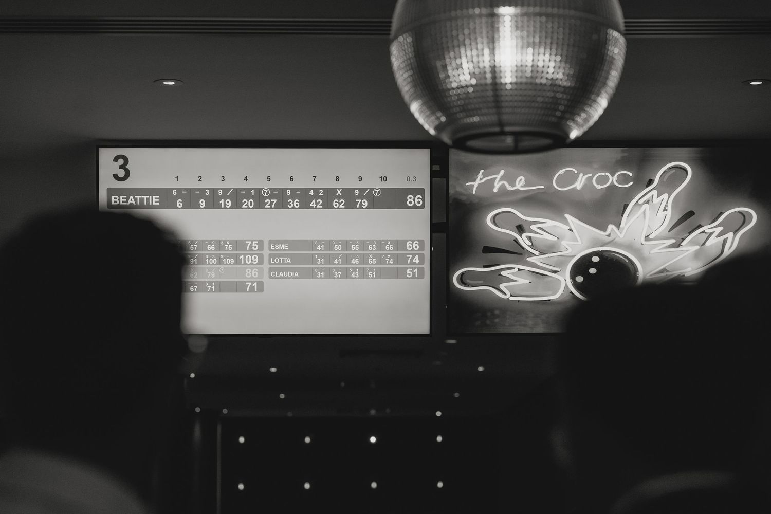 A black and white photo of a bowling alley scoreboard with a disco ball and neon sign.