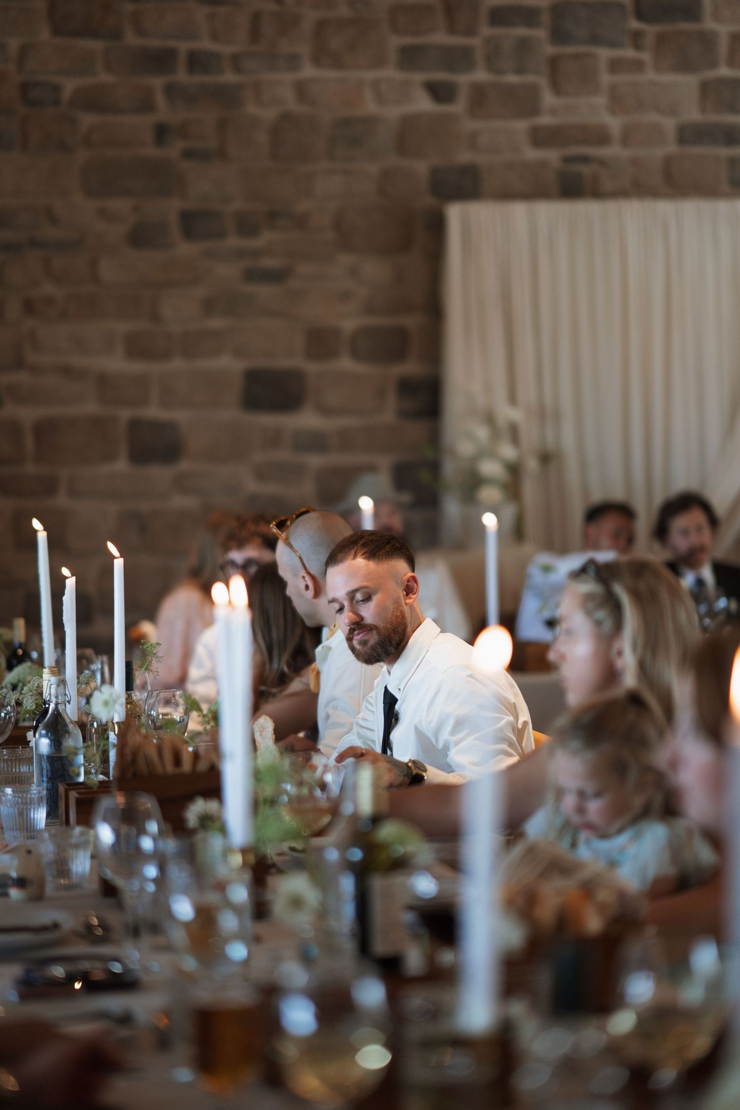 Wedding guests seated at a long table with candles and decor against a rustic brick wall.