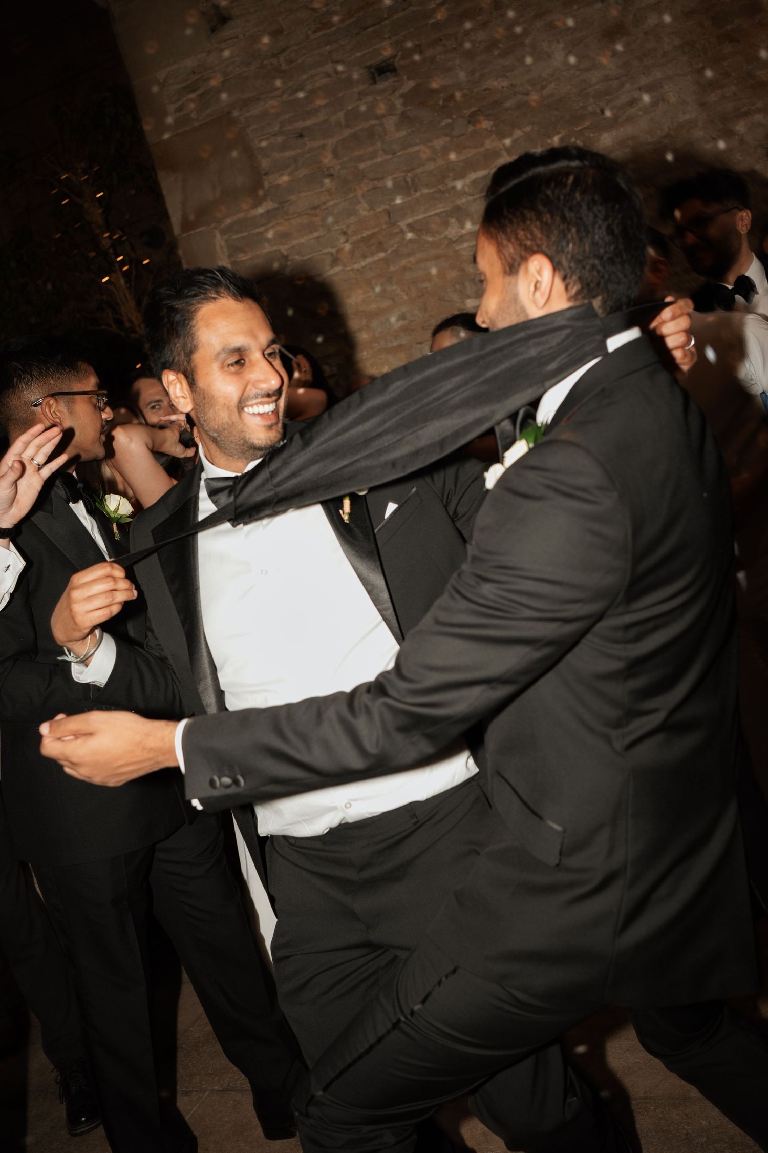 Wedding guests in black tuxedos dancing and embracing on the dance floor.