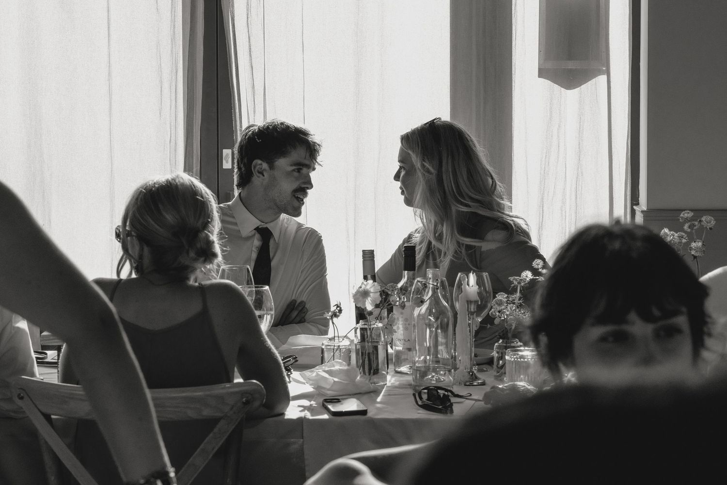 Wedding reception table setting with guests visible in silhouette against bright windows.